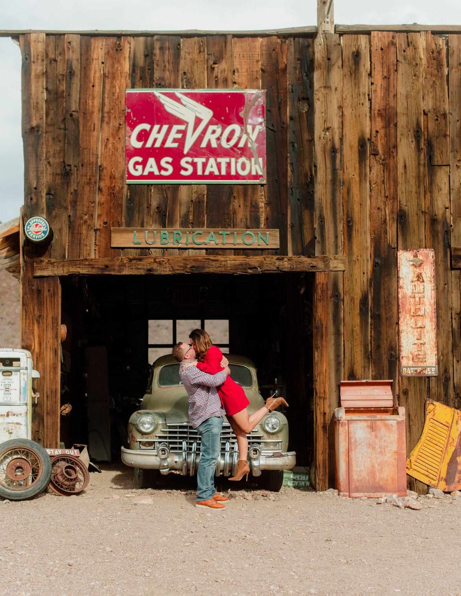 Couple kissing under a rustic Chevron Gas Station sign with a vintage car in the background.
