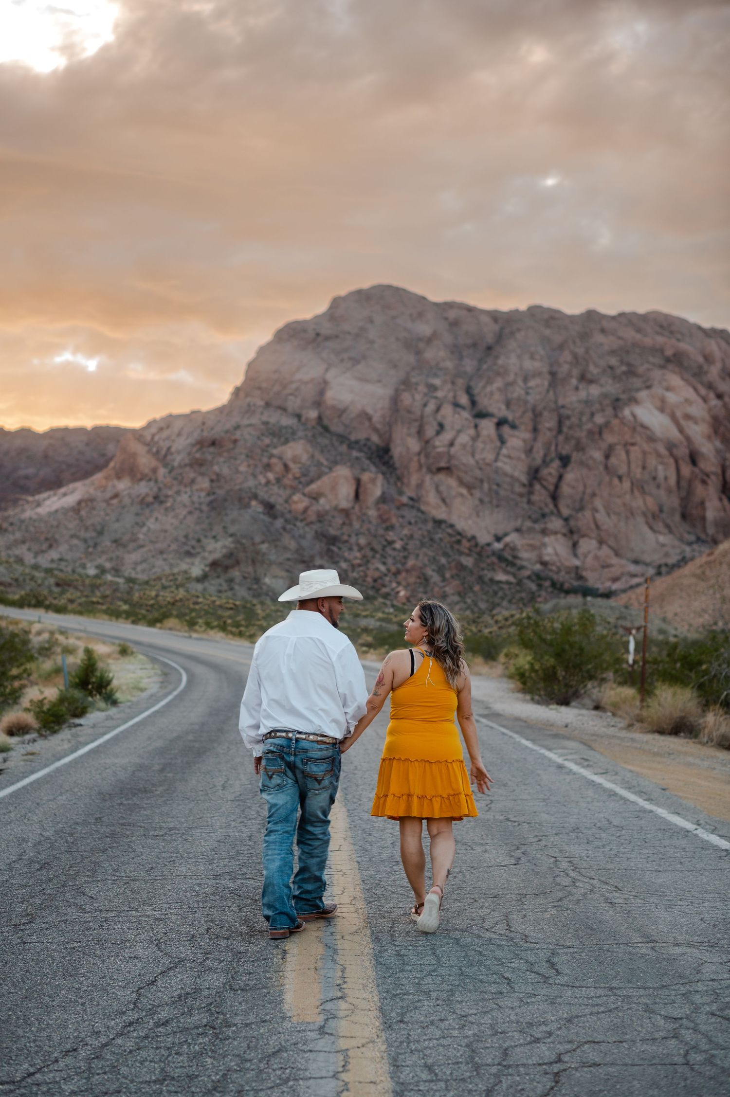 Couple walks hand-in-hand down an empty desert highway at sunset with rocky mountains ahead.