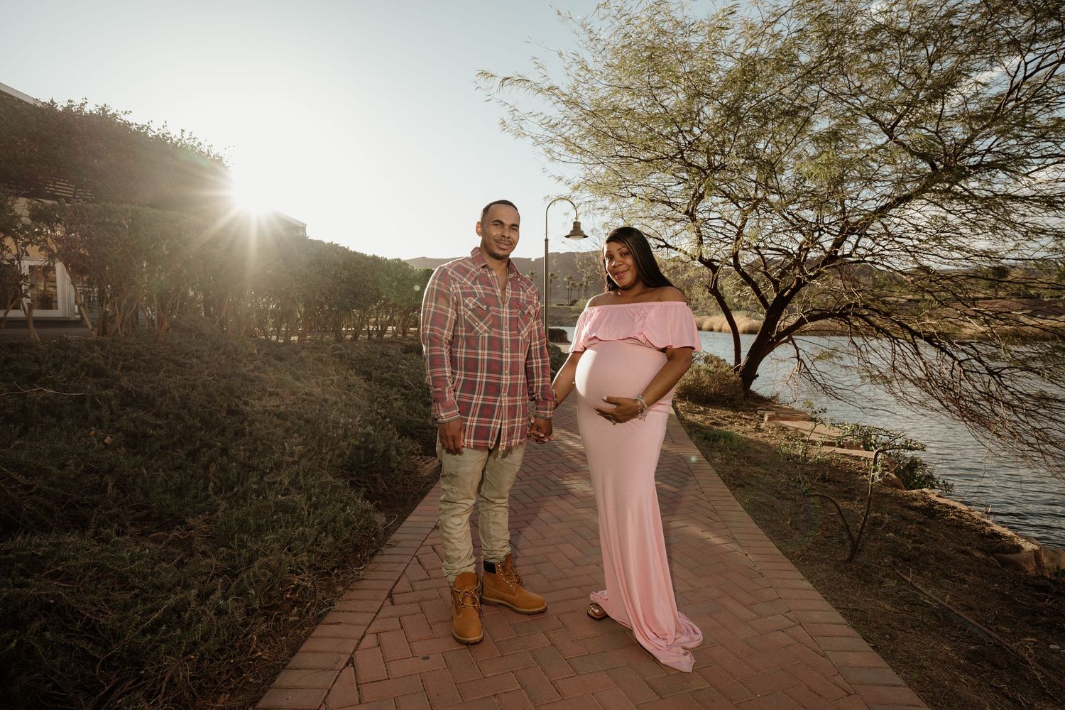 Couple standing on sunlit brick walkway, pregnant woman in flowing pink maternity dress beside her partner.