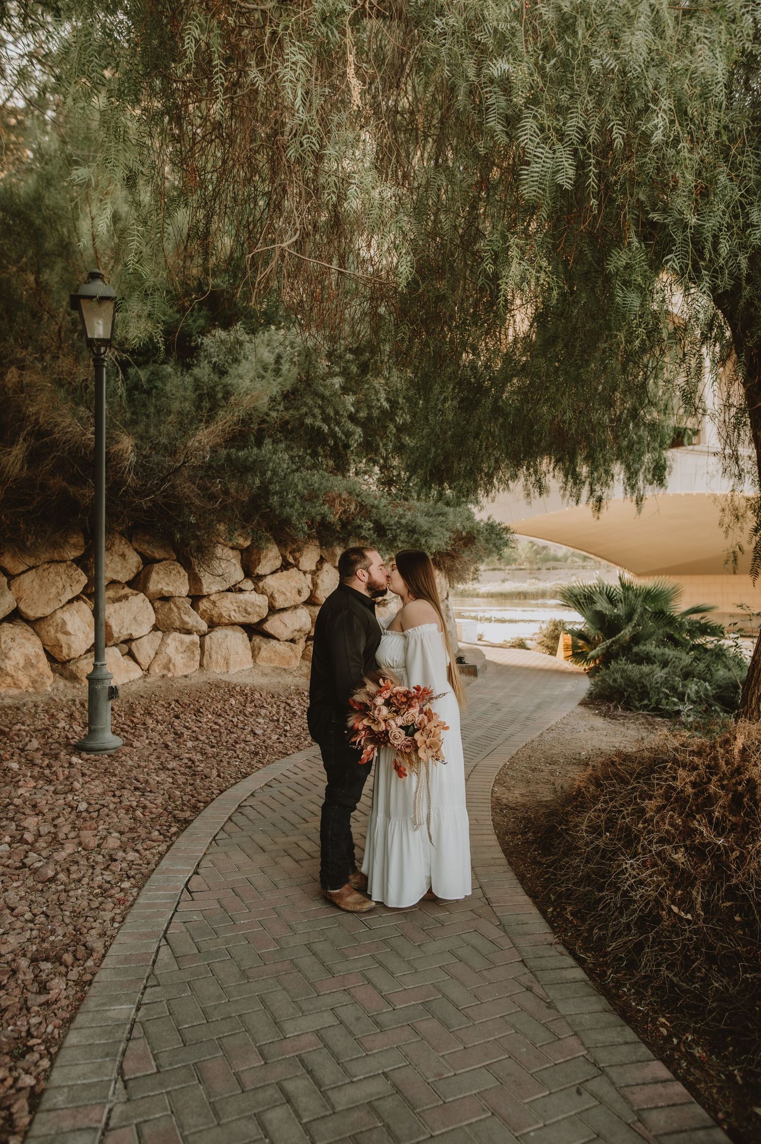 Bride and groom sharing romantic kiss under weeping willow tree on brick path.