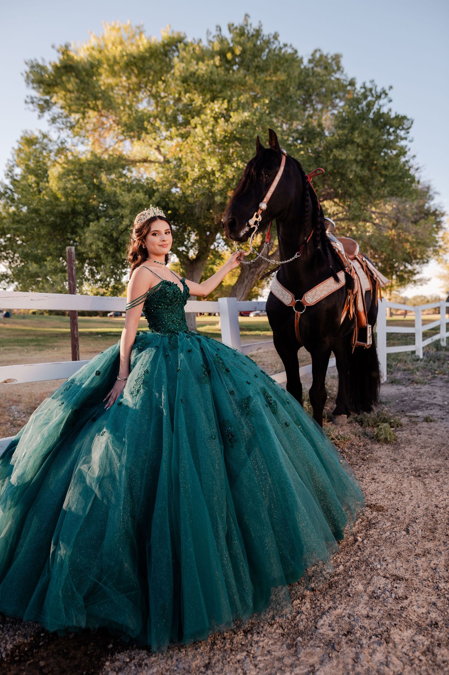 Elegant quinceañera in teal tulle ball gown standing beside a saddled dark horse at sunset.
