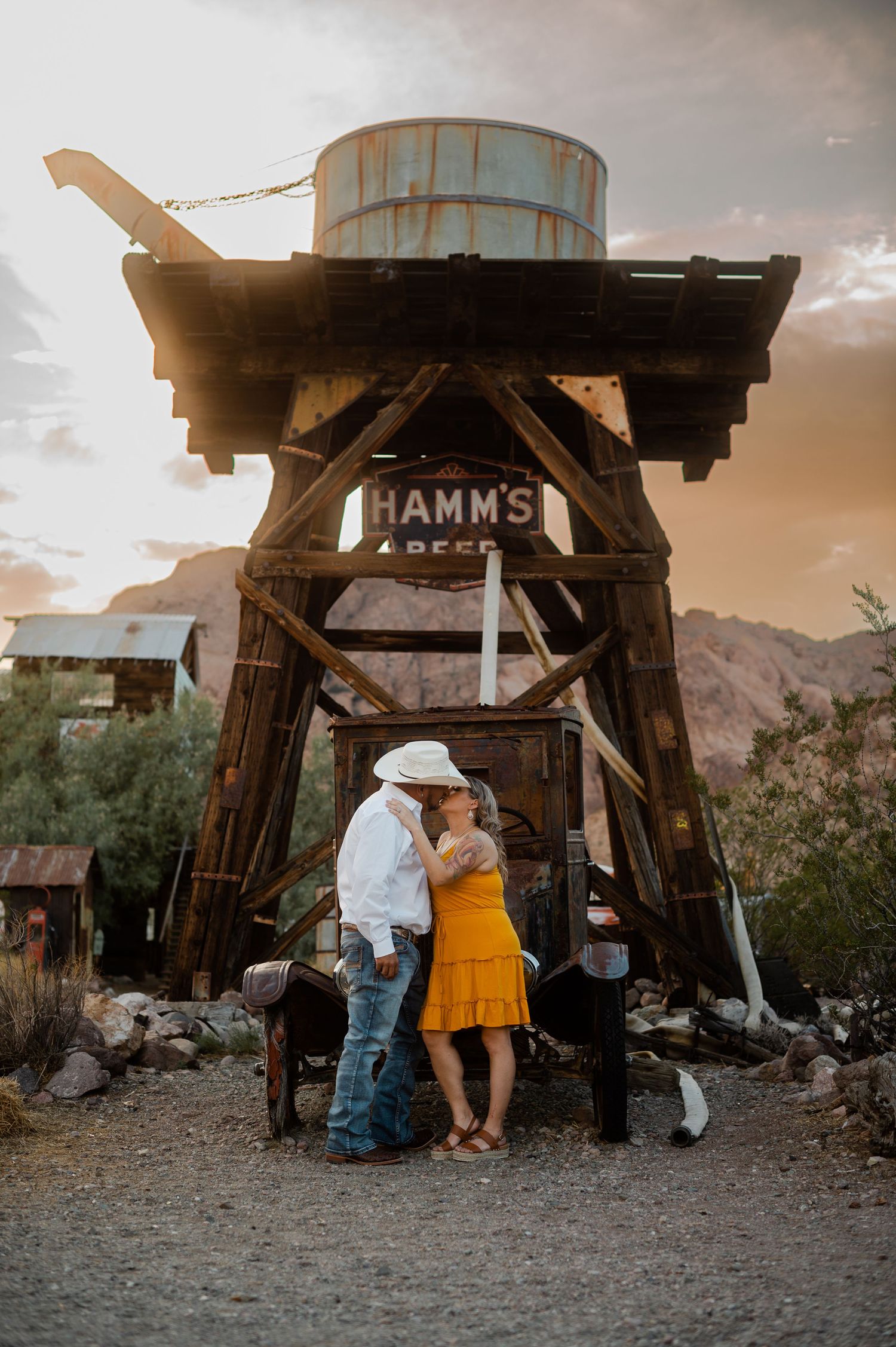 Couple kissing in front of a rustic Hamms Beer water tower at a desert ghost town.
