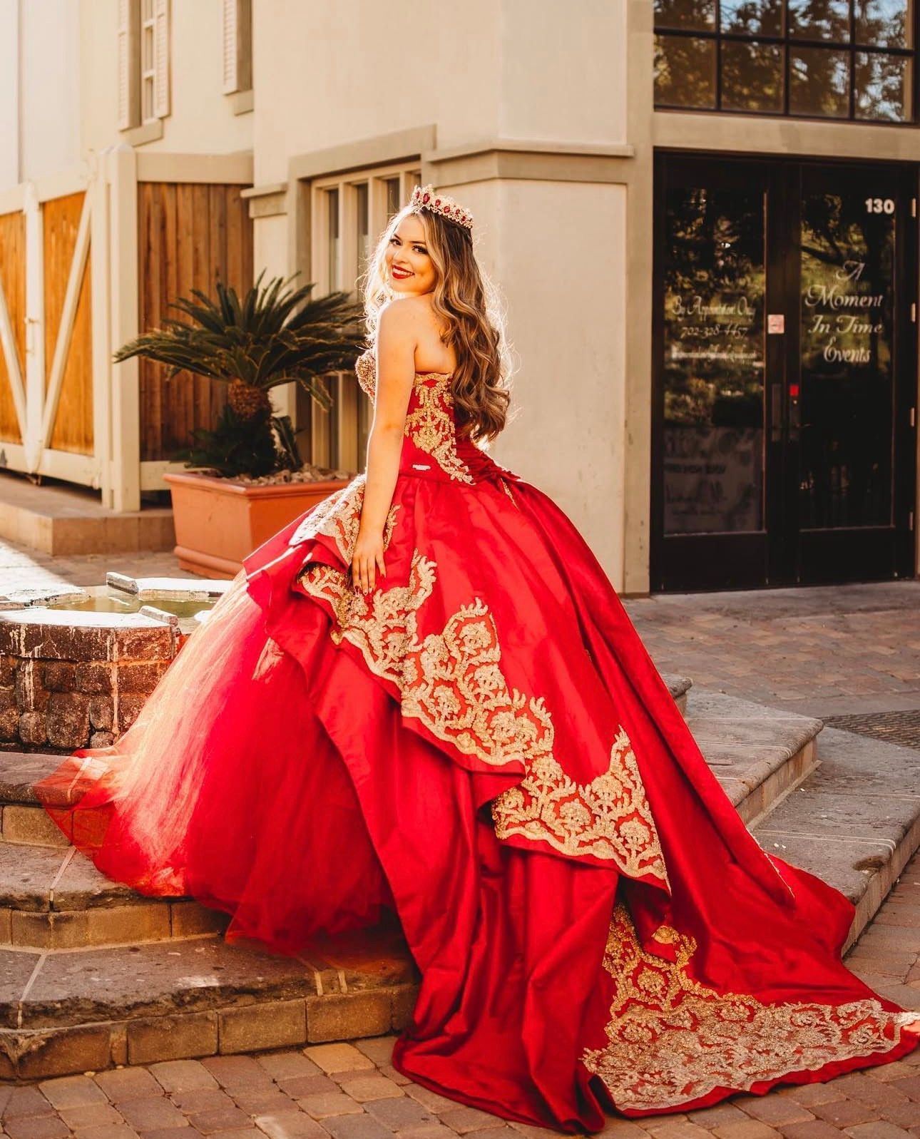 Young woman in red and gold quinceañera ball gown with crown posing on steps outside building.
