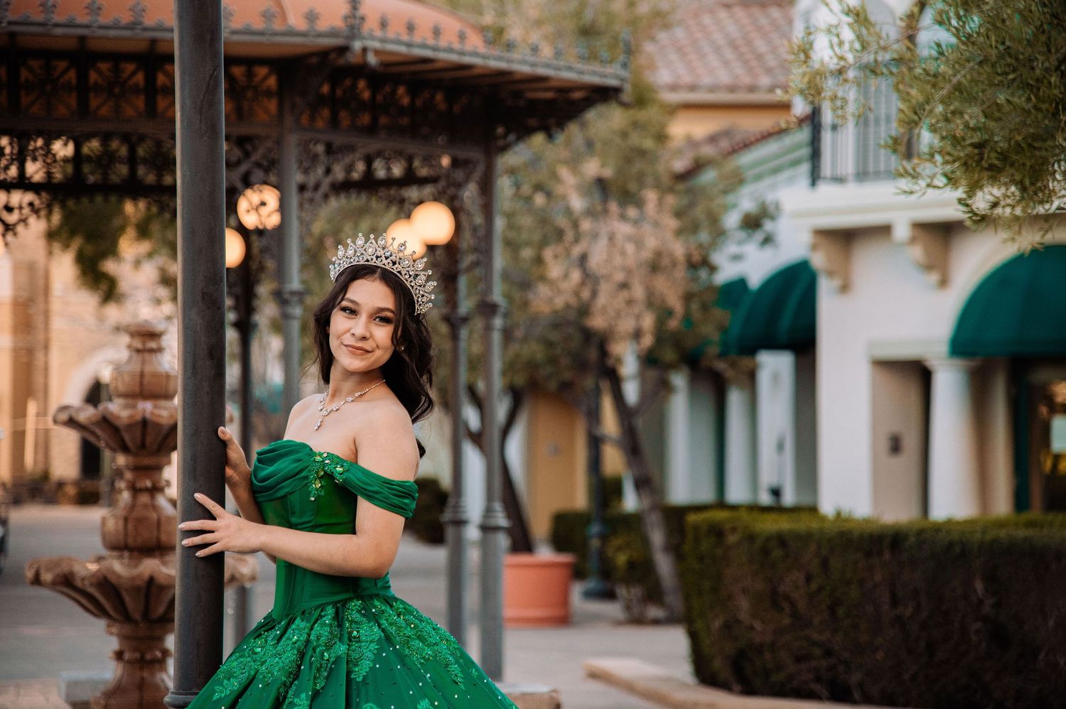 Young woman in emerald green quinceañera ball gown and crown posing by a lamp post at an outdoor venue.