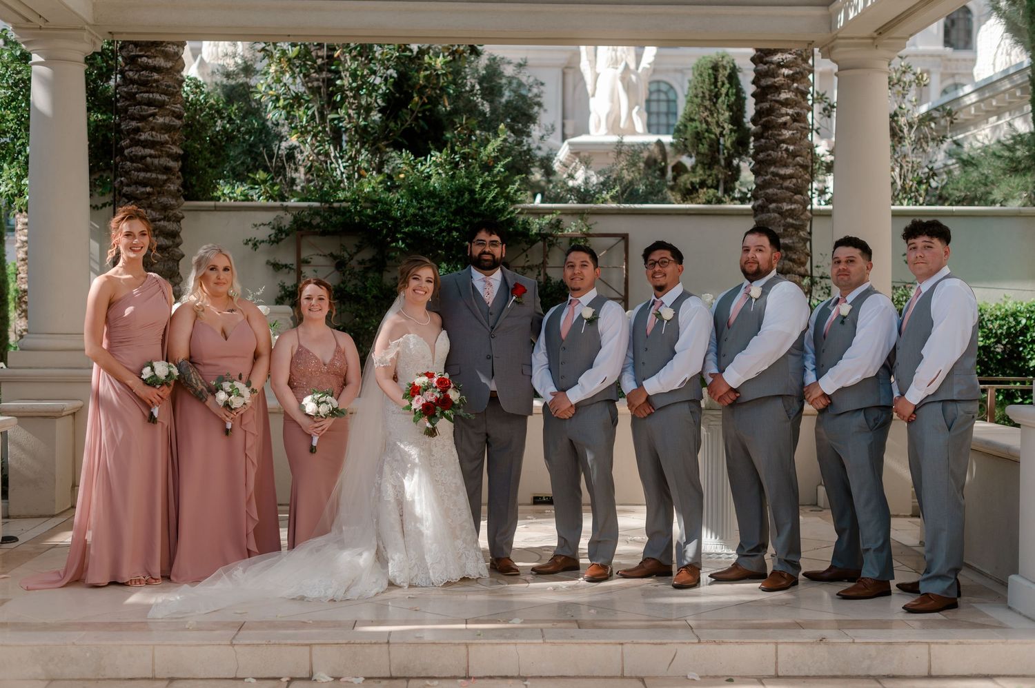 Wedding party poses in dusty rose bridesmaid dresses and gray suits at an elegant outdoor venue.