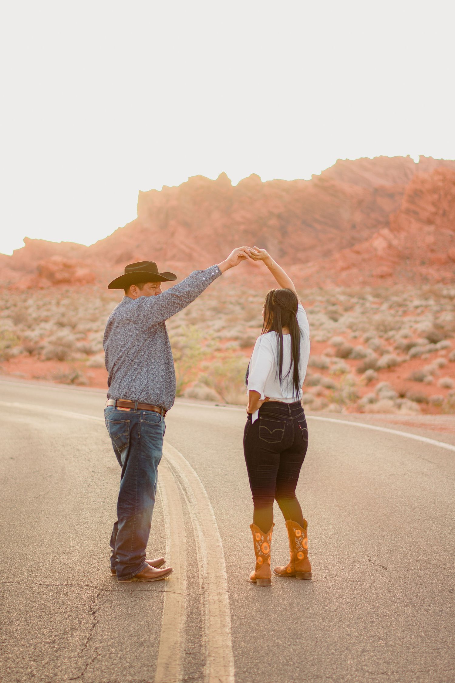 Couple dances on empty desert highway at sunset with red rock formations glowing in the background.