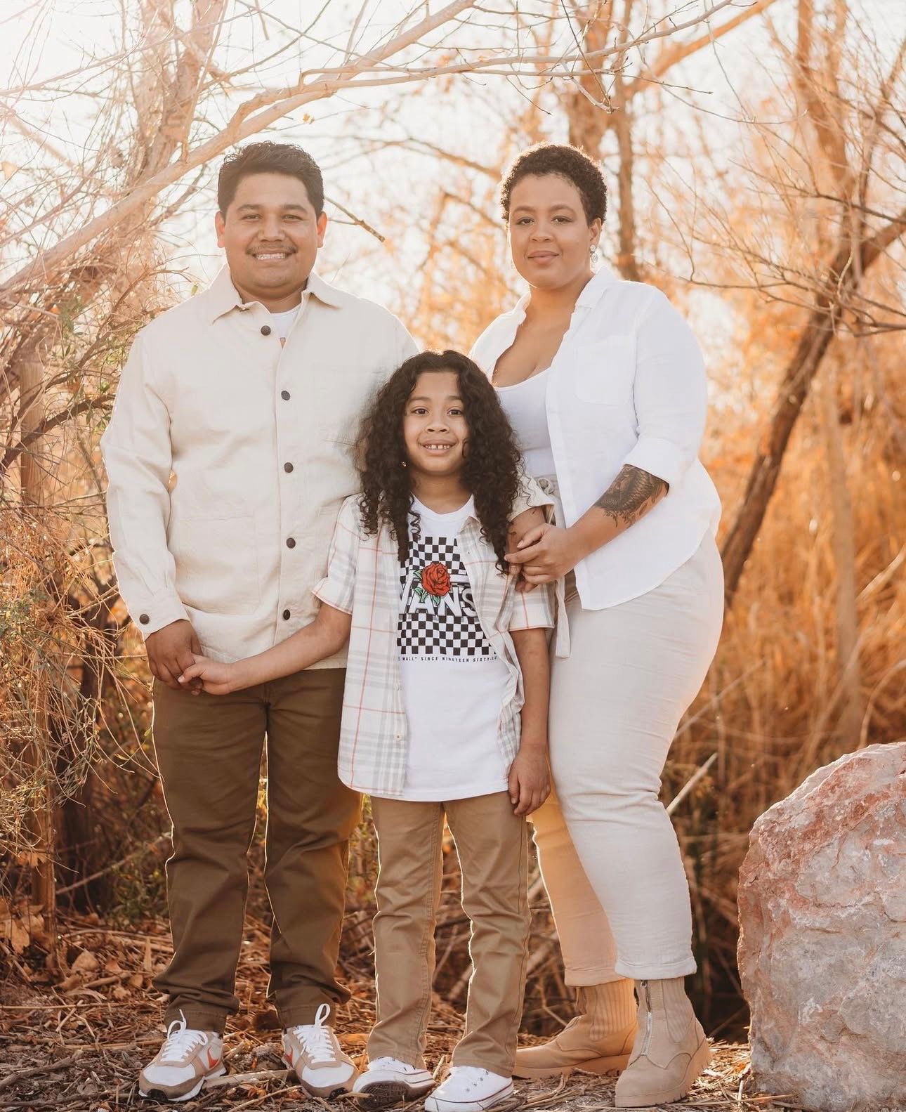 A family of three poses together outdoors among autumn trees, dressed in coordinating neutral tones.