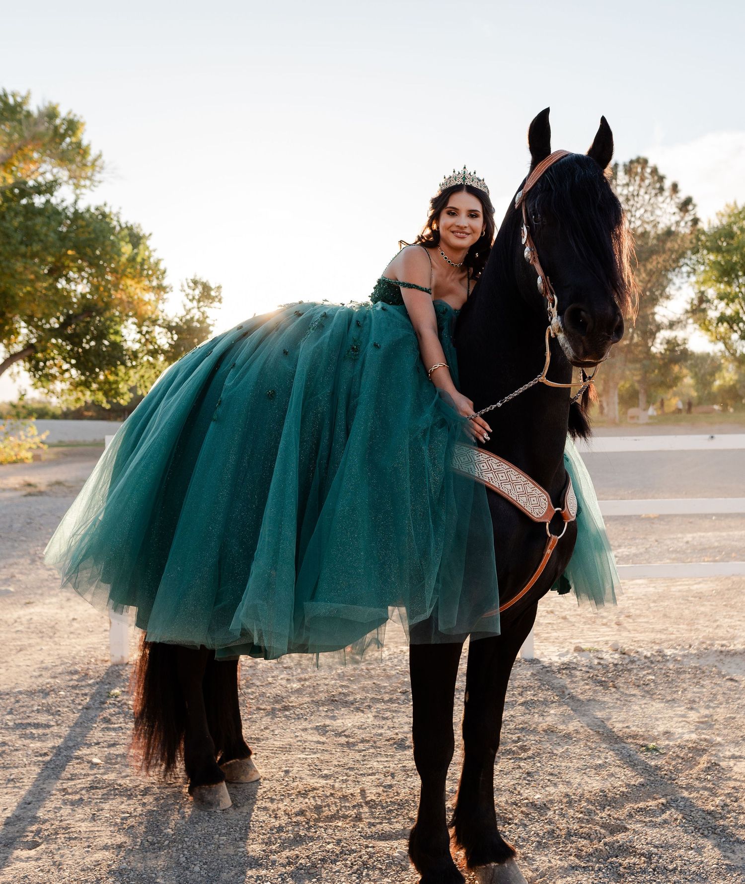 Smiling young woman in teal quinceañera ball gown and crown poses with a black horse at dusk.