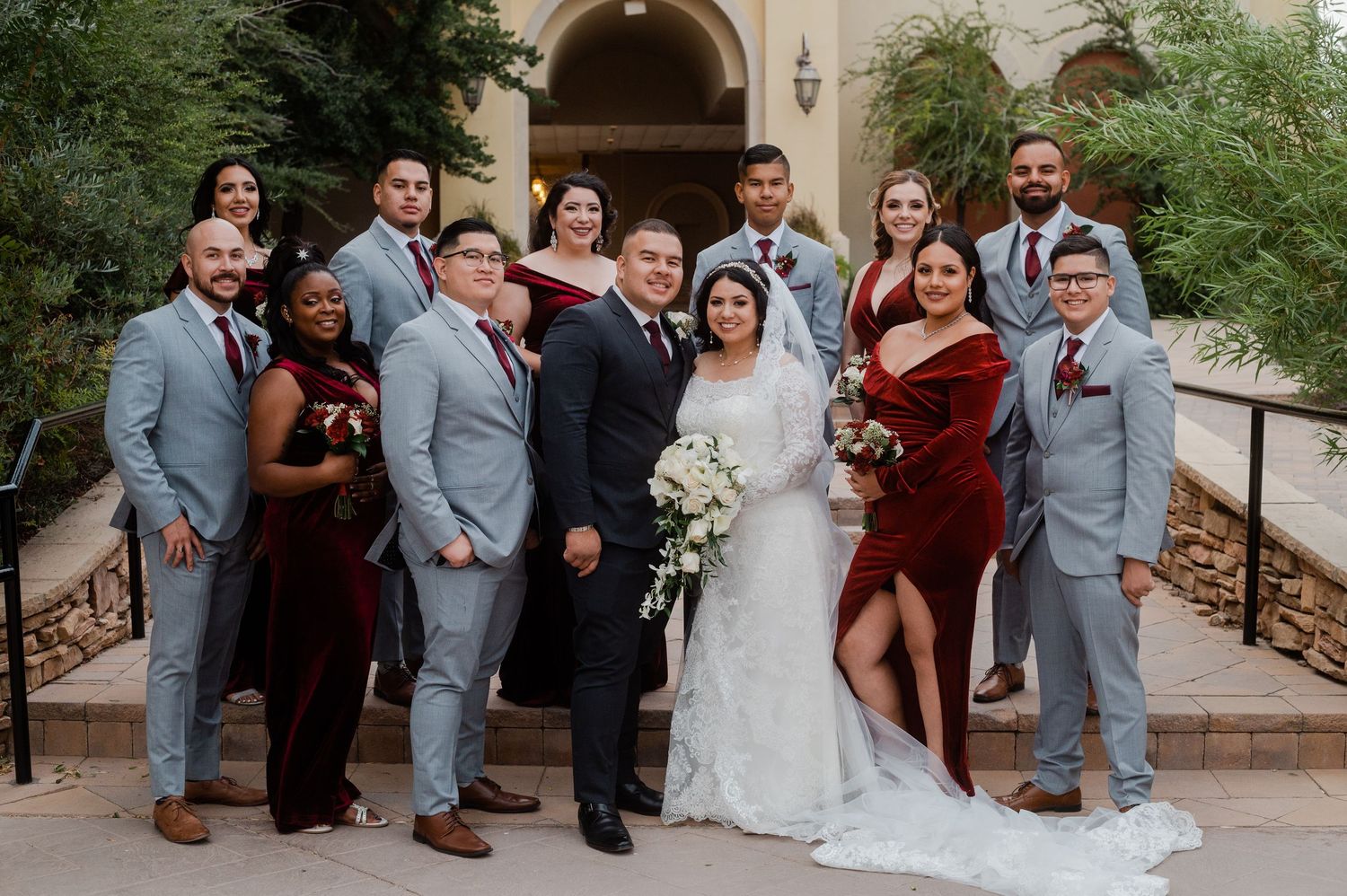 Wedding party in burgundy and gray poses on steps outside a southwestern-style wedding venue.
