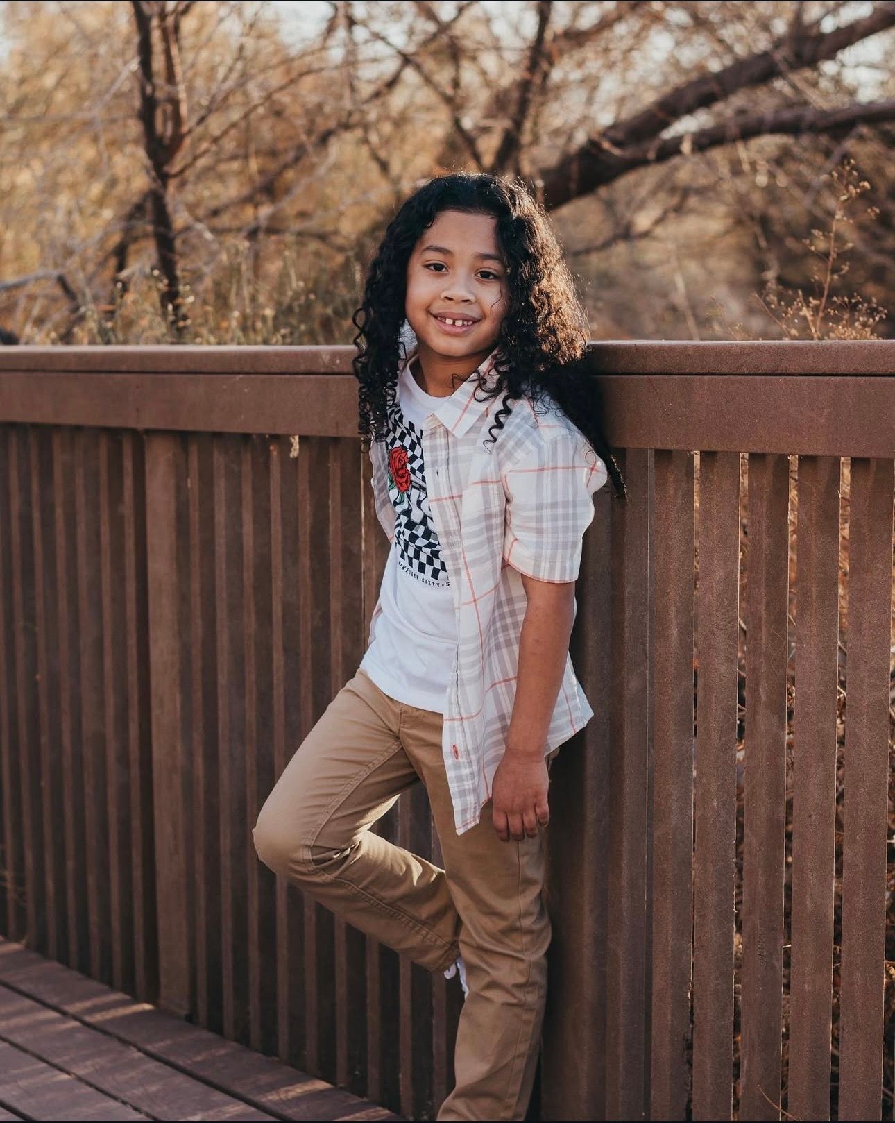 A smiling child with curly hair leans against a wooden fence wearing a plaid shirt and khaki pants outdoors.