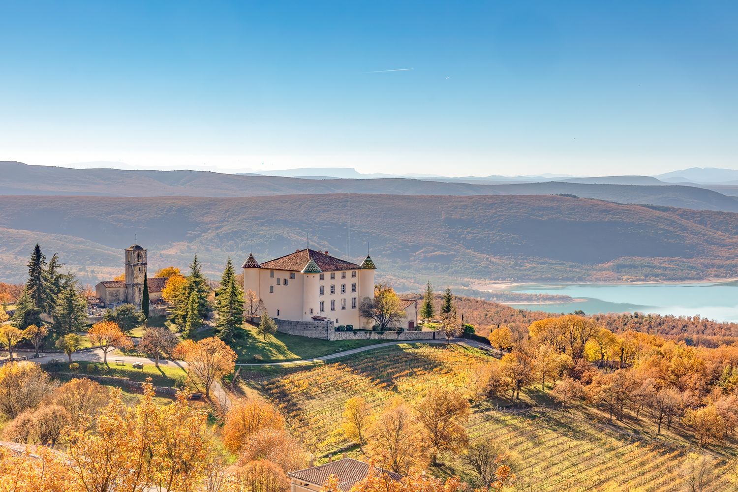 Aerial view of a historic castle surrounded by autumn vineyards with a serene lake and mountains in the background.