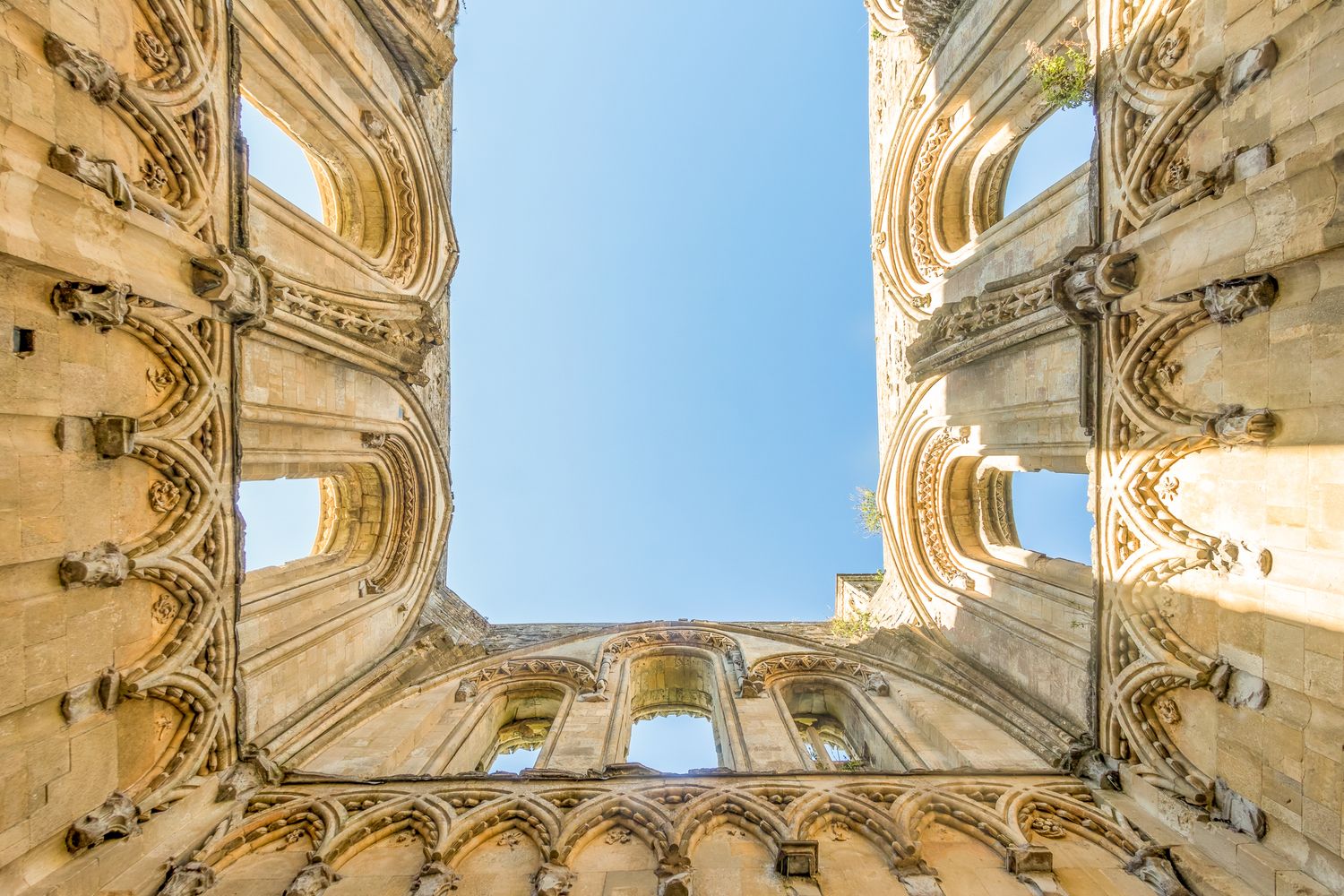 Upward view of ornate Gothic stone ruins framing a clear blue sky with intricate architectural details.