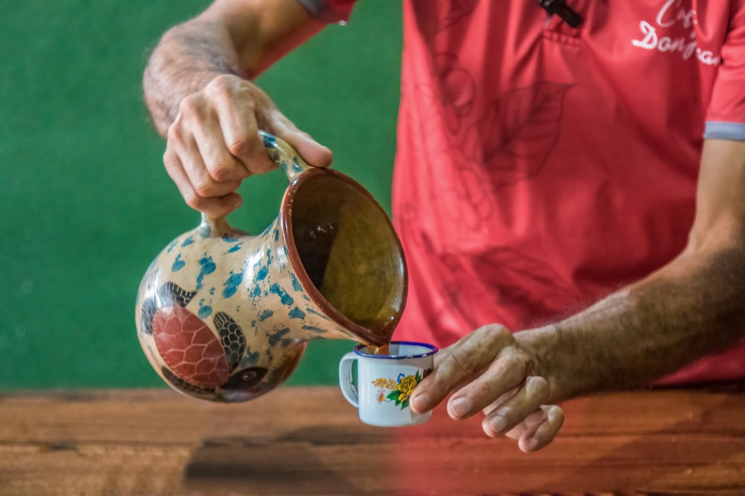 Man in red shirt pouring coffee from decorative ceramic pitcher into a small mug on a wooden table.