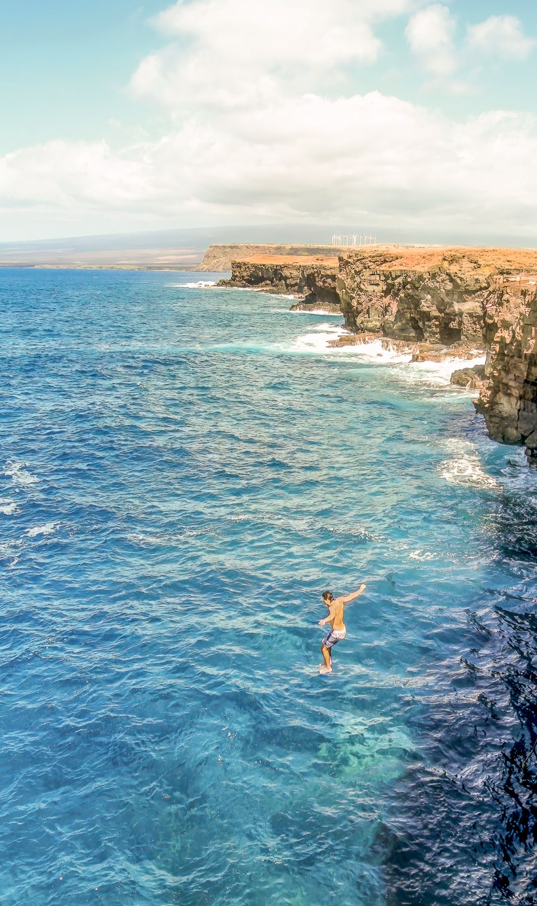 Person cliff jumping into vivid blue ocean waters beside dramatic volcanic rock formations under a partly cloudy sky.
