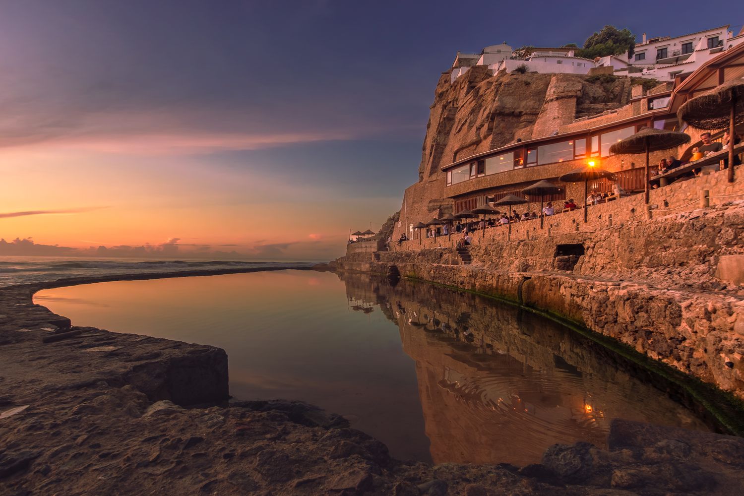 Stunning sunset over a coastal cliff village with warm golden reflections shimmering in a calm tidal pool below.