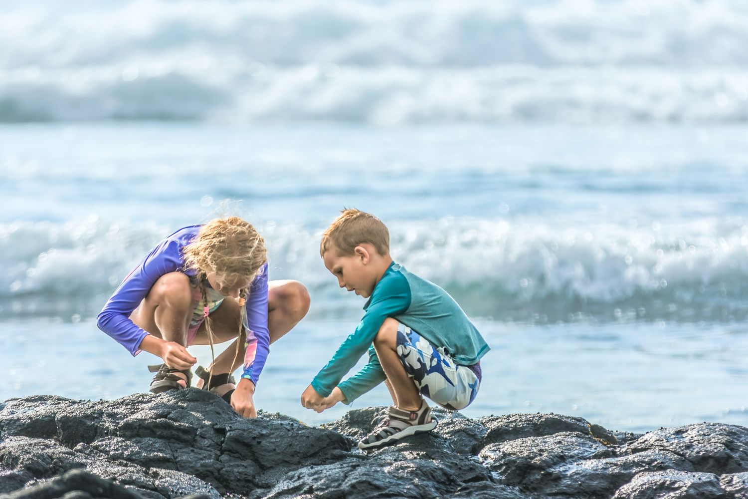 Two children explore tide pools on rocky volcanic shore with ocean waves in the background.