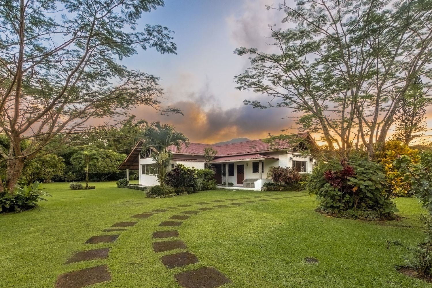Tropical home with lush green lawn, stone path, and colorful sunset sky surrounded by tall trees and tropical gardens.