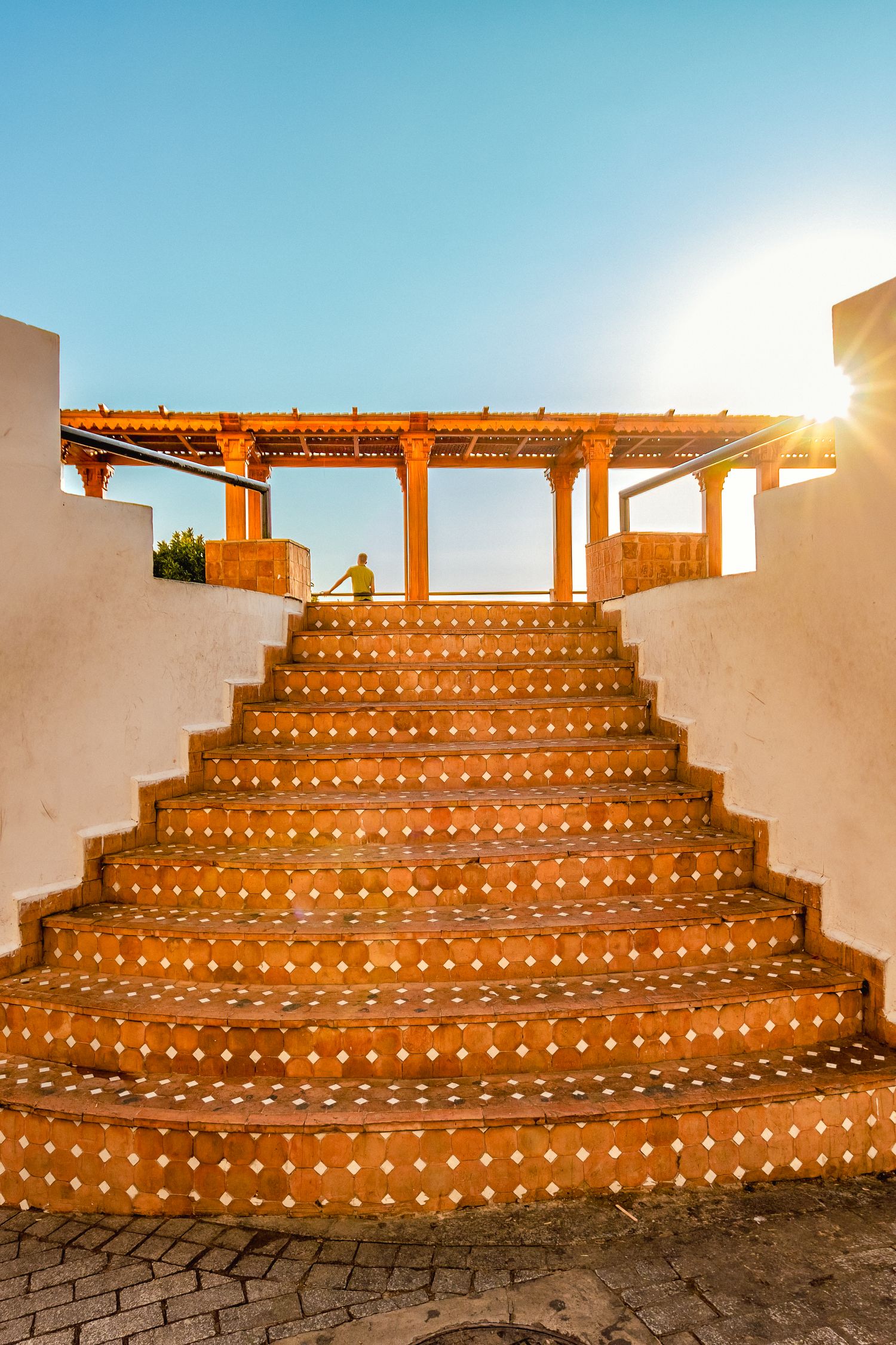 Ornate terracotta tiled staircase leading to a wooden pergola at sunset with warm golden sunlight.