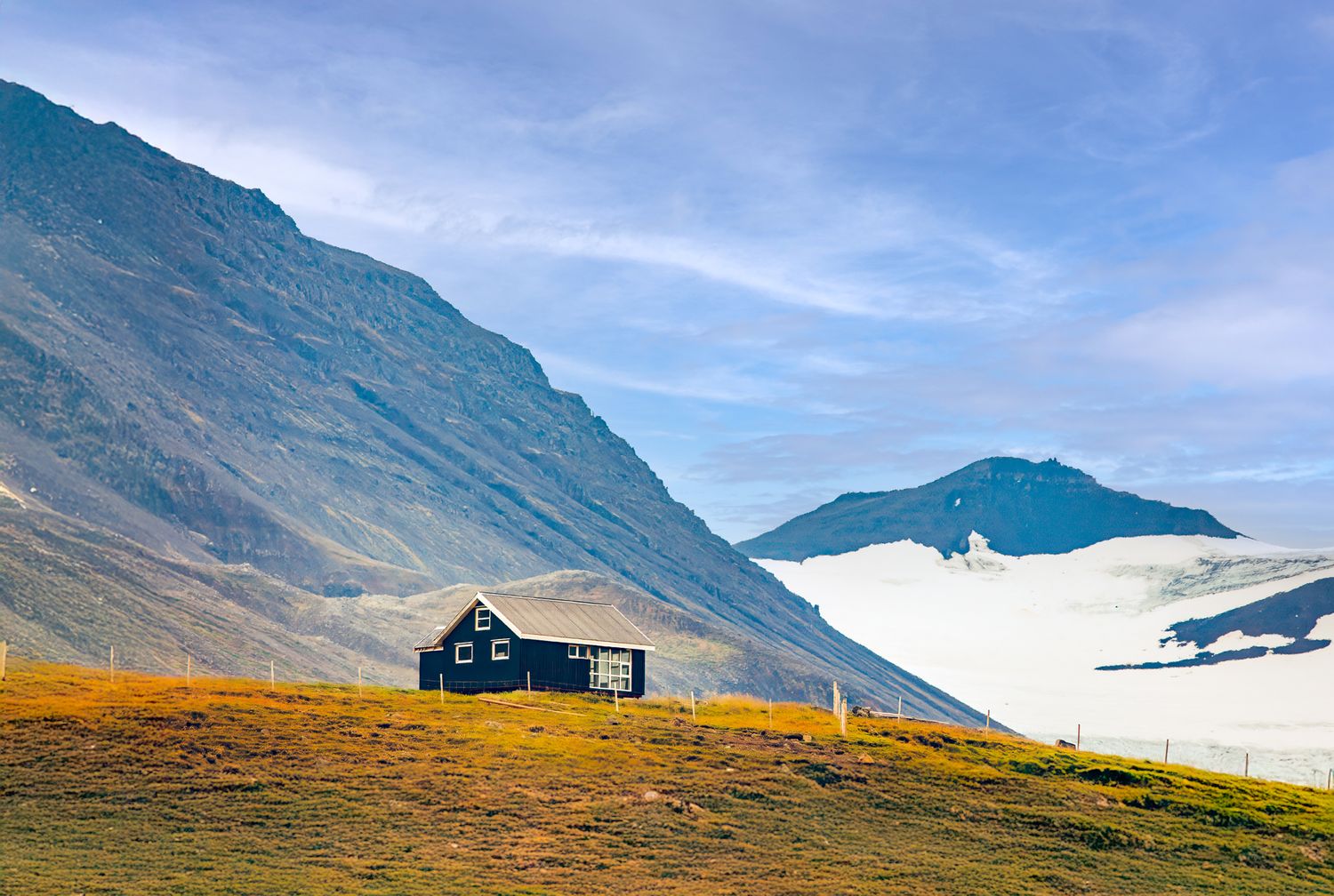 Remote black cabin on golden hillside with dramatic mountain glacier backdrop under clear blue sky in Iceland.