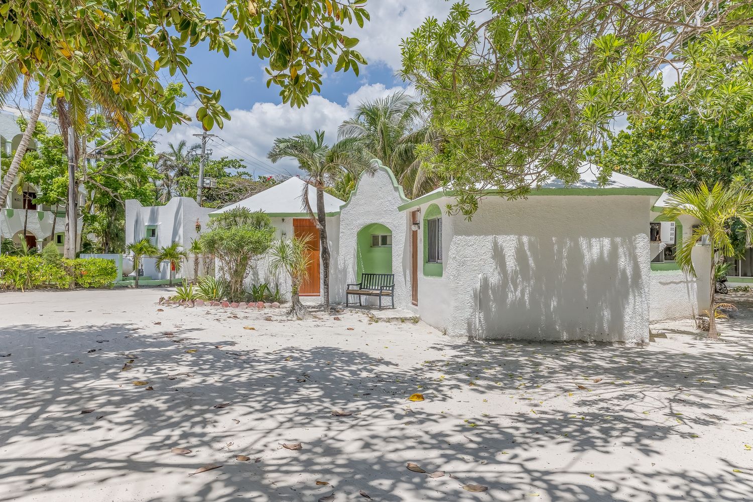 White stucco cottage with green arched door, surrounded by tropical trees and sandy yard in a sunny Caribbean setting.
