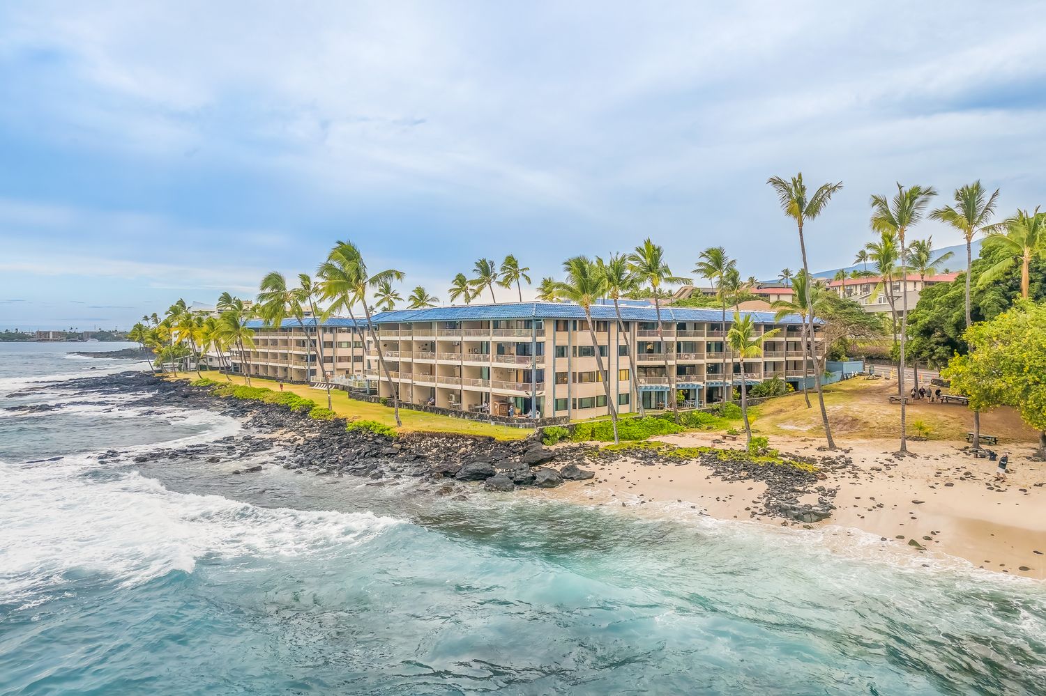Aerial view of a beachfront hotel with blue balconies, palm trees, and rocky shoreline in Hawaii.