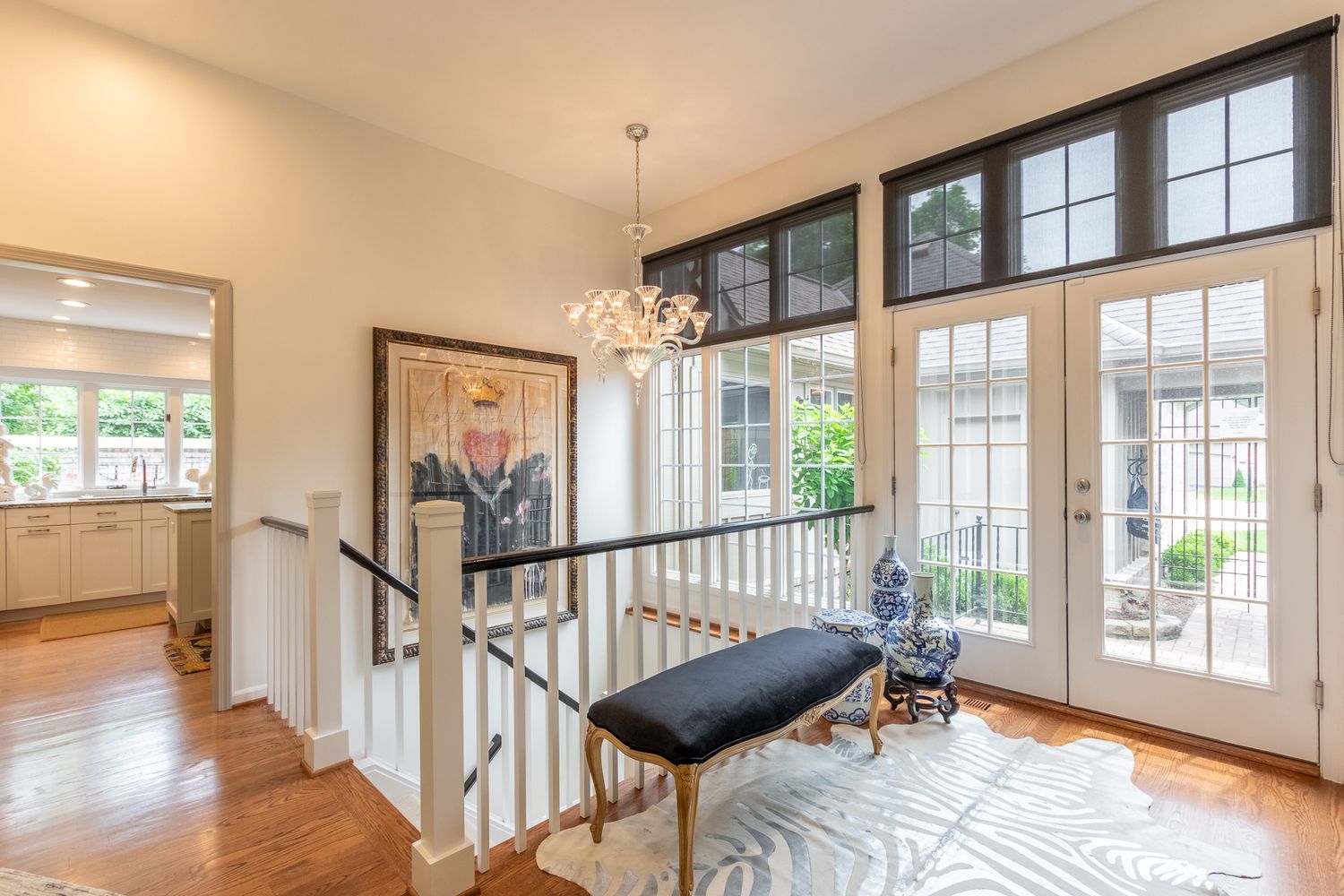 Bright upper landing with hardwood floors, chandelier, French doors, and transom windows overlooking greenery.