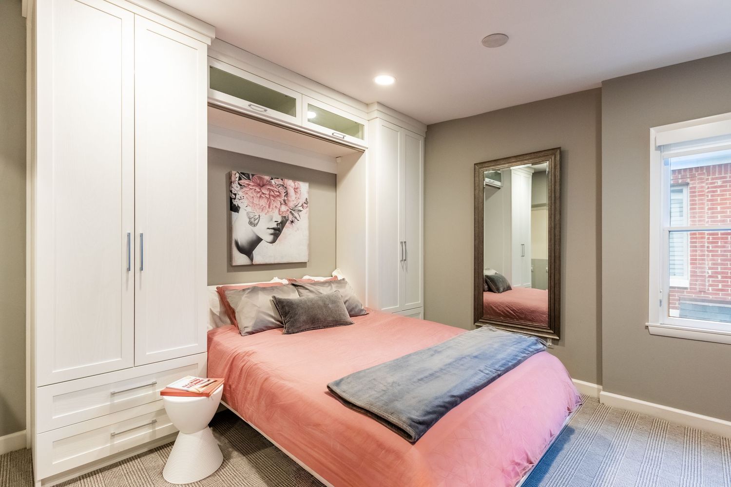Cozy bedroom with built-in white Murphy bed, pink bedding, large mirror, and gray walls in a compact space.