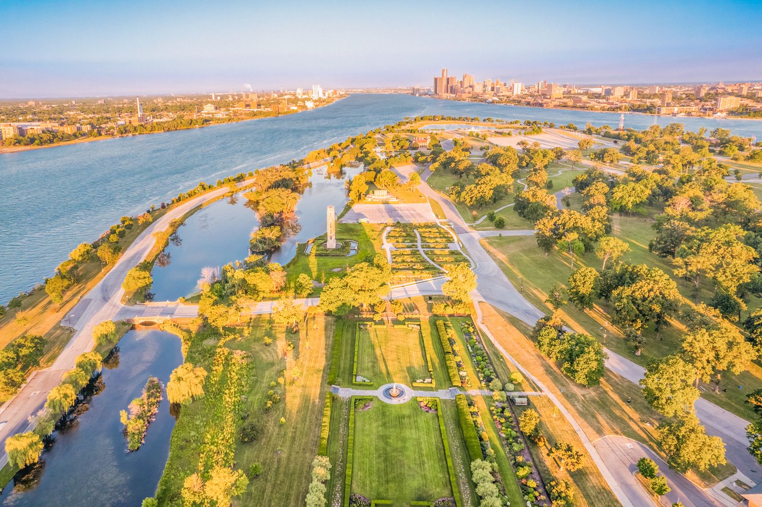 Aerial view of riverside park with formal gardens, pathways, and city skyline at golden hour.