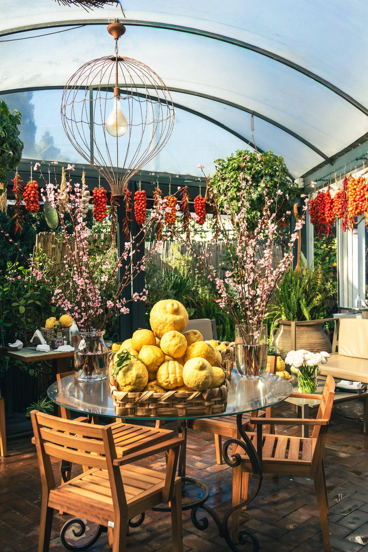 A charming greenhouse dining area with wooden chairs, a glass table topped with yellow gourds, hanging red peppers, and lush greenery.
