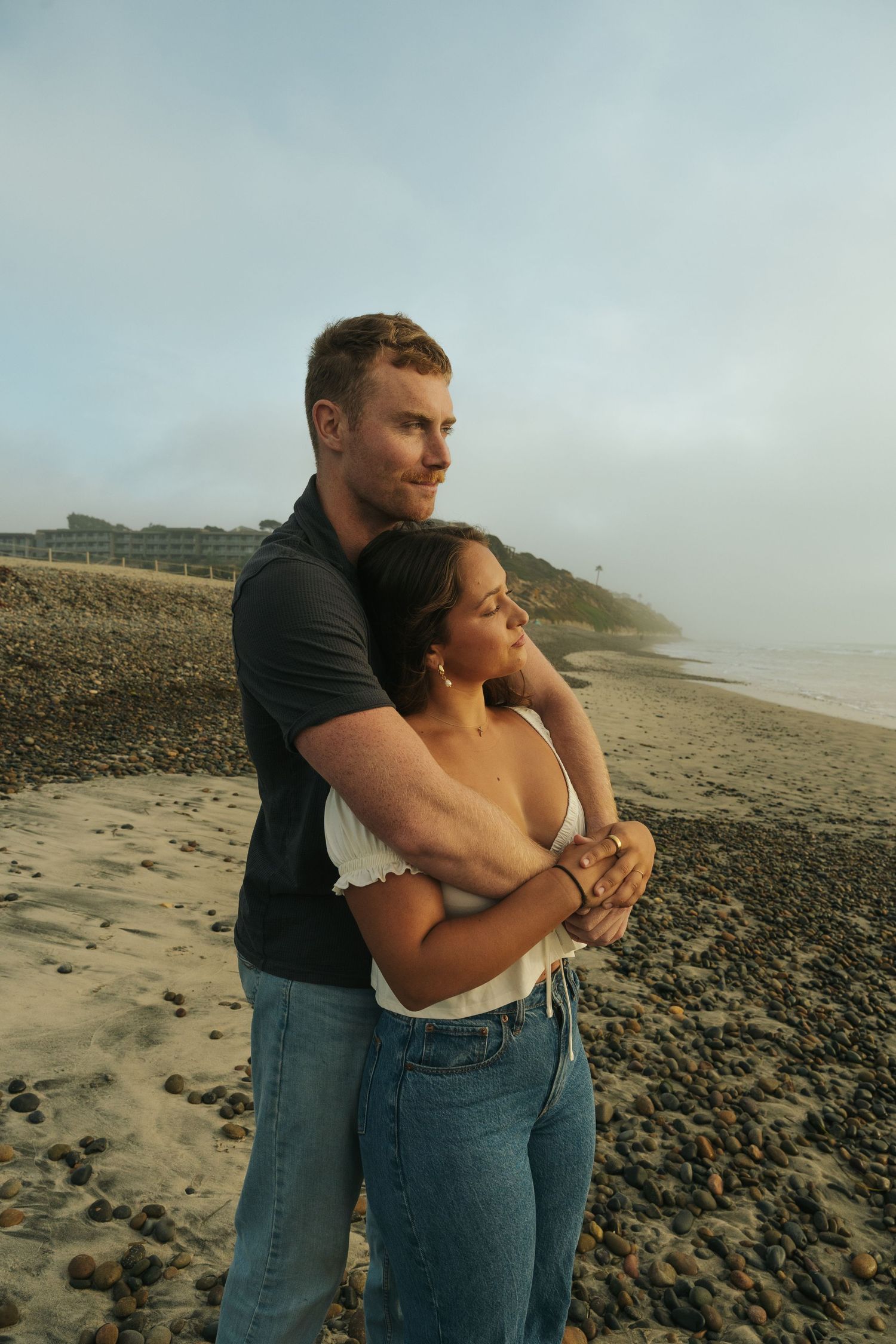 Couple embracing on a rocky beach with coastal cliffs in the background.