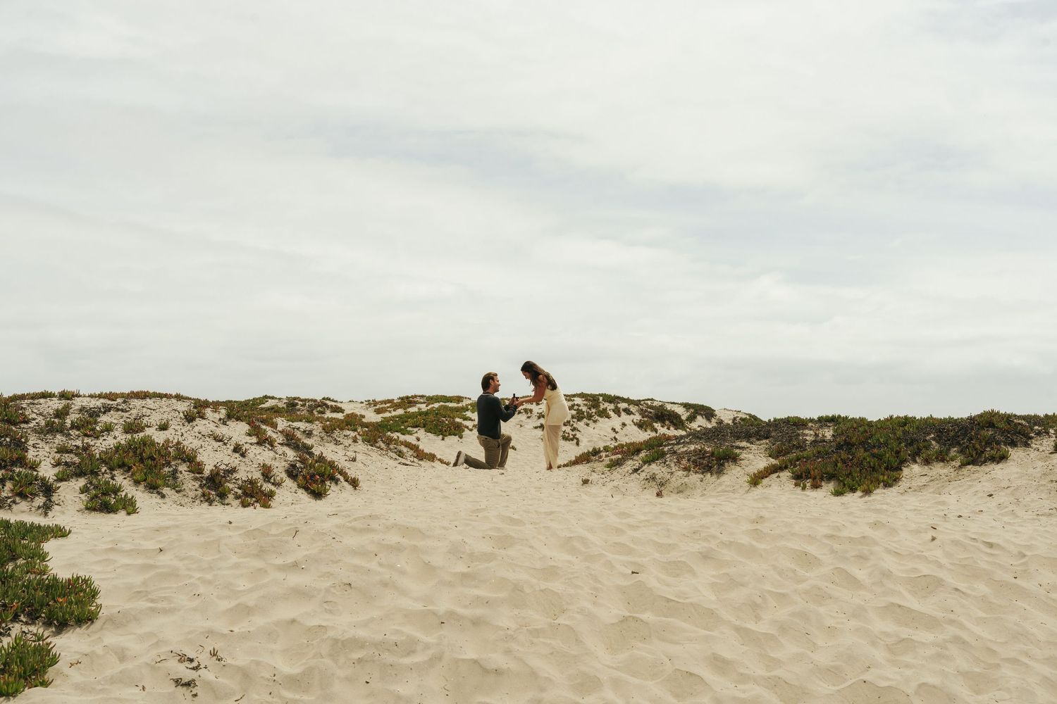 A person proposing on sandy beach dunes under overcast skies, with sparse coastal vegetation in the background.