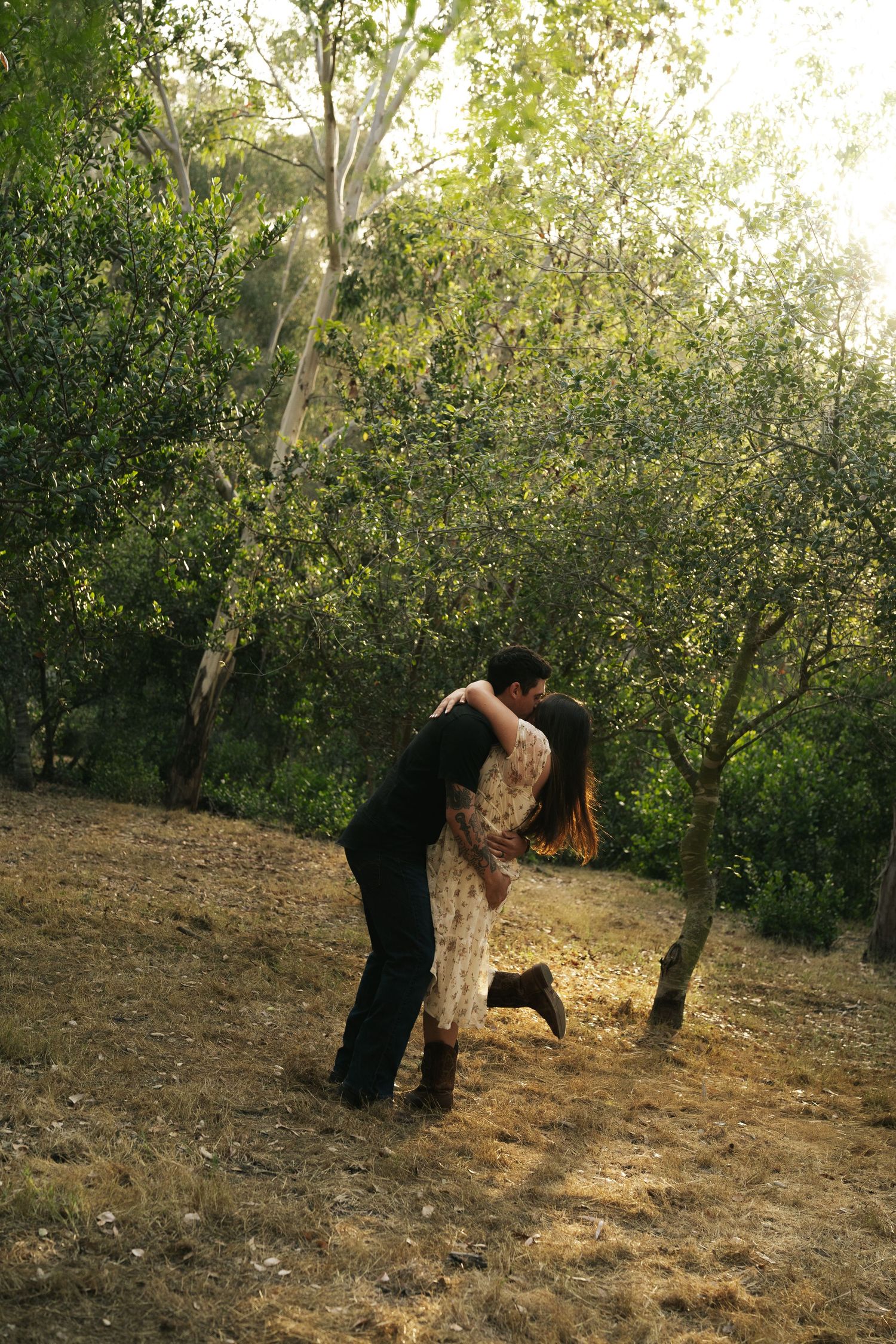 Man lifting woman in a joyful embrace among sunlit trees in a woodland setting.
