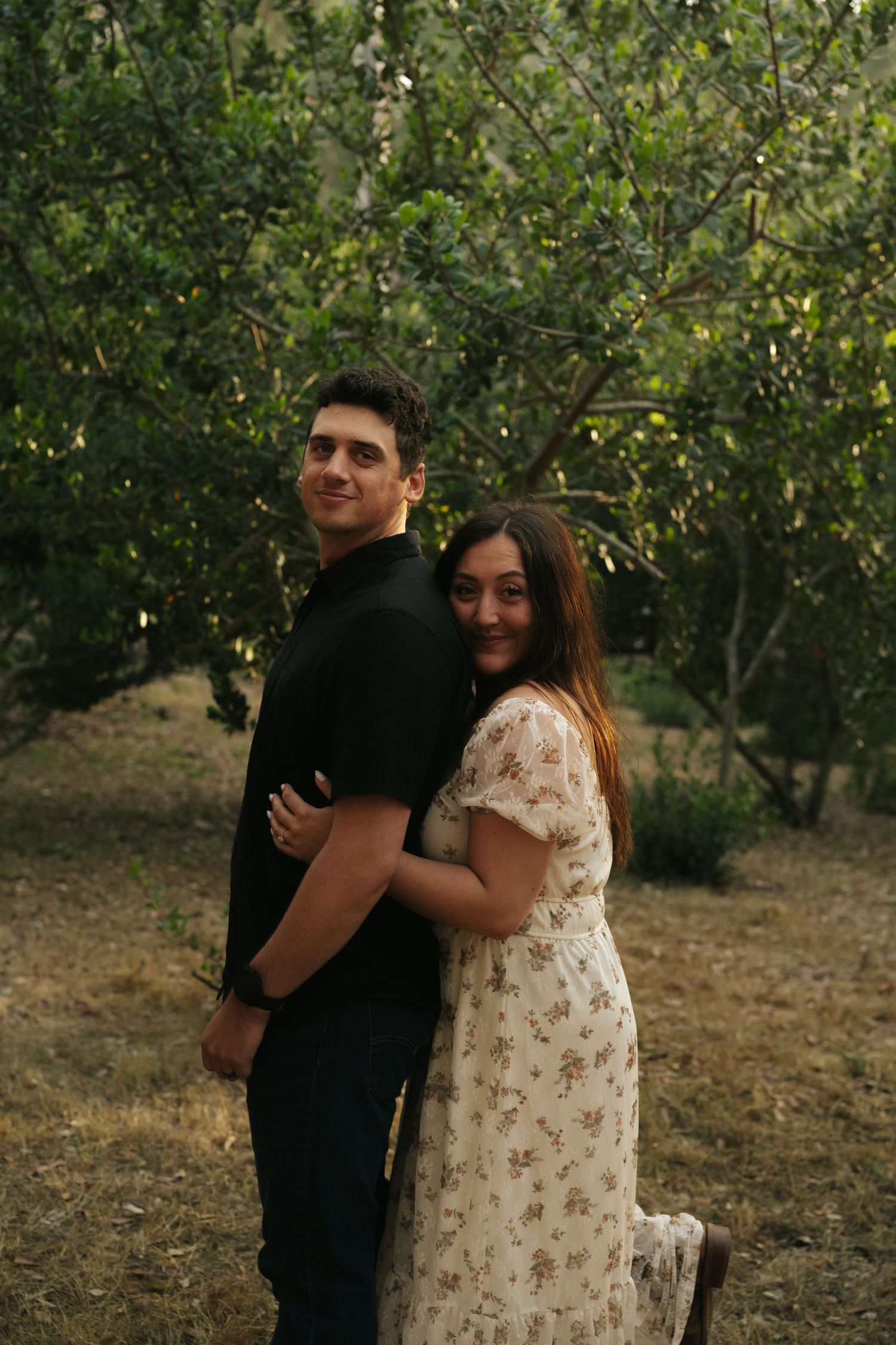 Couple posing back-to-back with woman hugging man from behind in a lush green orchard.