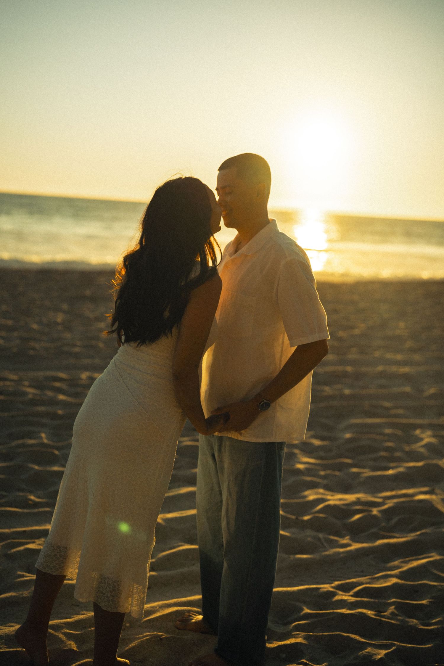 A couple shares a romantic kiss on a sandy beach at golden sunset, with ocean waves glimmering in the background.