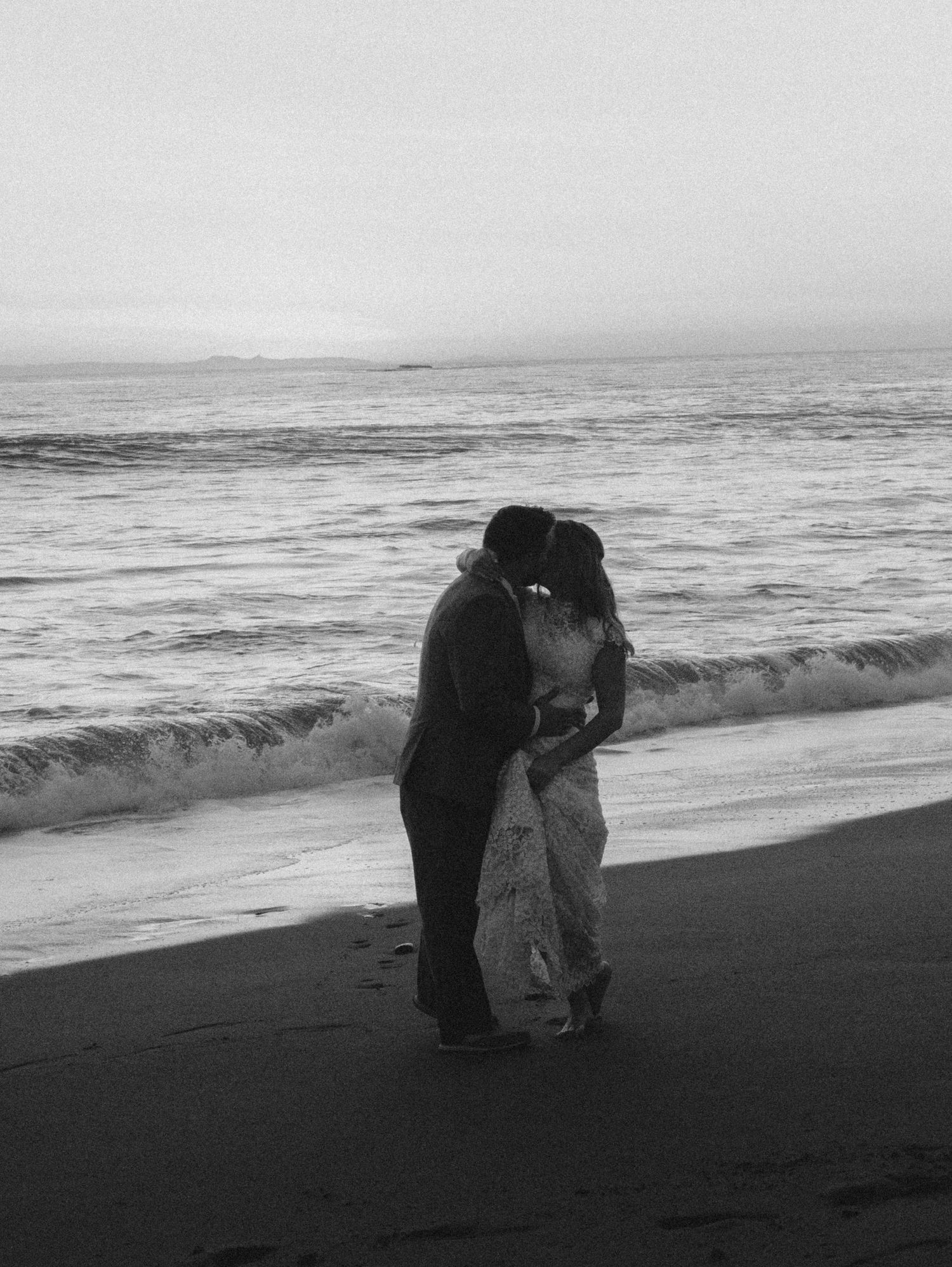 Black and white photo of newlyweds kissing at the water's edge as gentle waves wash ashore.