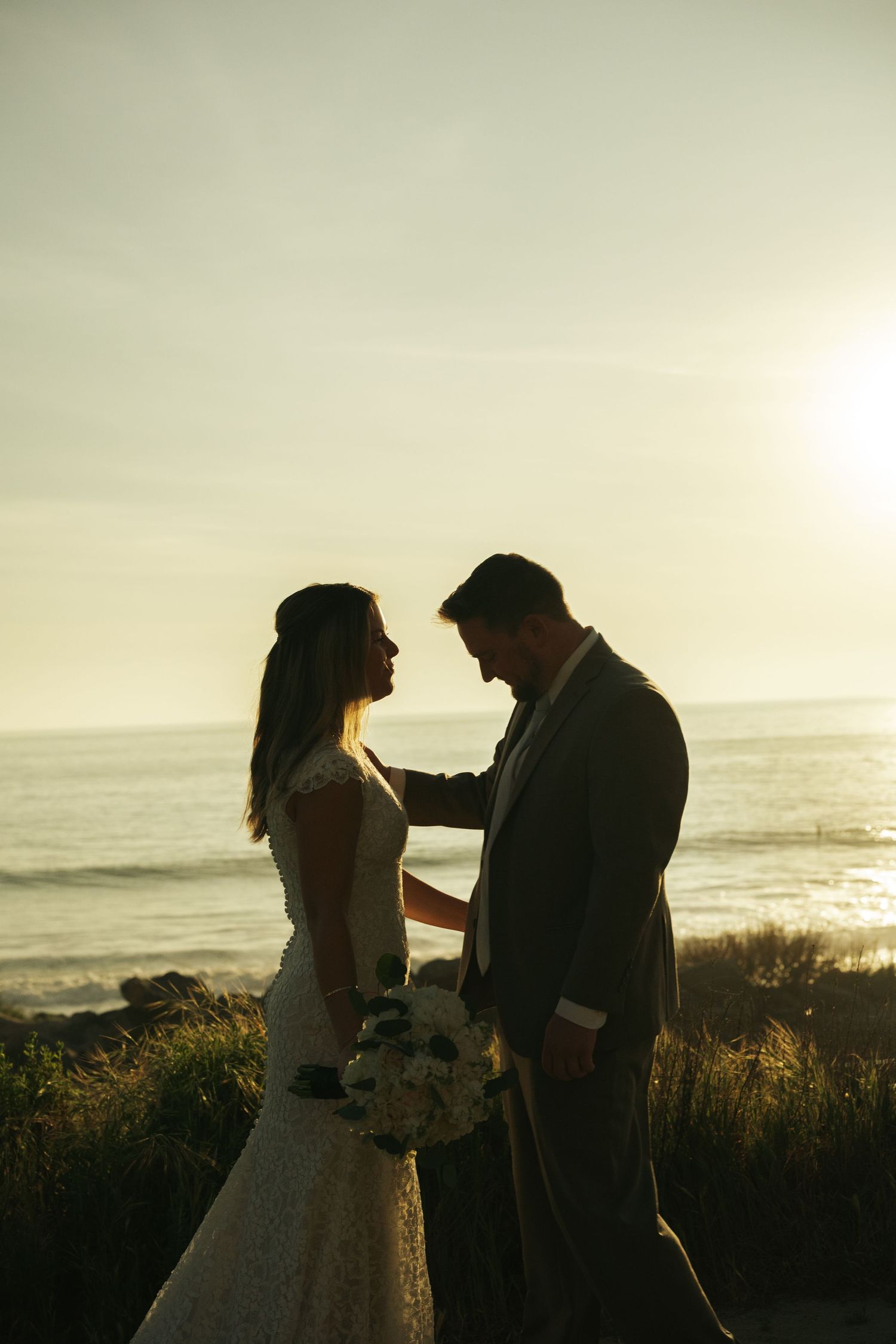 Bride and groom silhouetted against golden sunset at ocean cliffside, holding bouquet of white flowers.