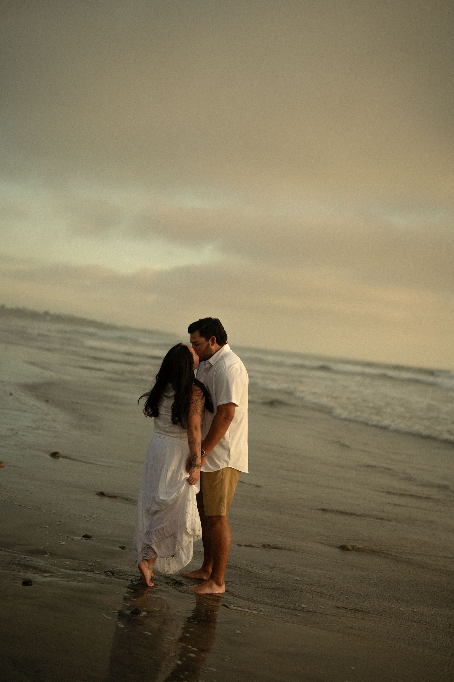 Couple sharing a romantic kiss on a serene beach at dusk with calm ocean waves behind them.