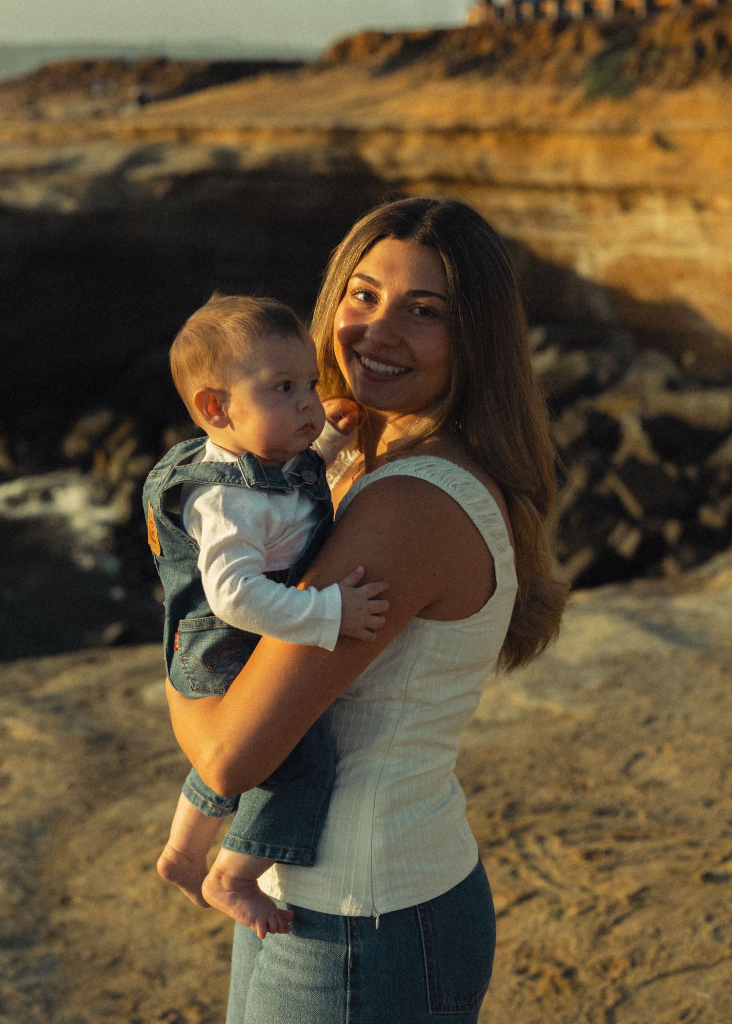 Smiling young mother holds her infant baby near rocky coastal cliffs in warm golden sunset light.