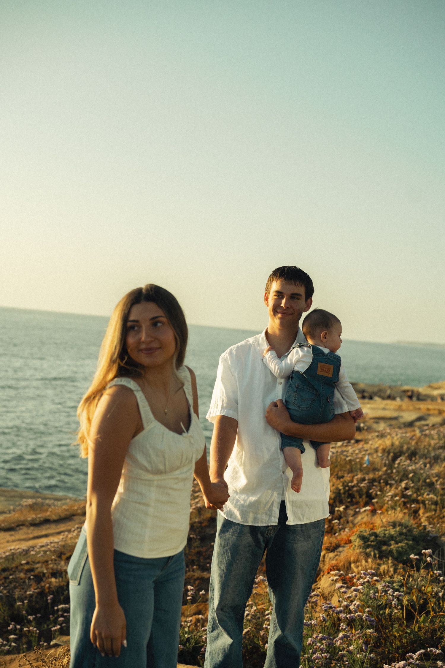Young family with baby posing on rocky coastal cliff at golden hour sunset.
