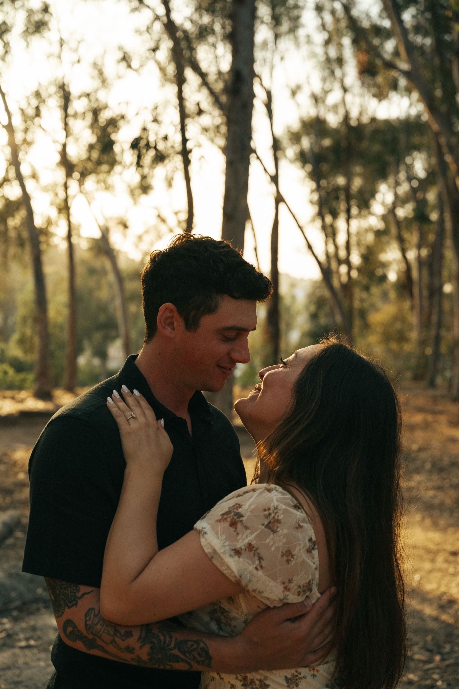 Couple leaning in for a kiss surrounded by tall trees in golden sunset light.