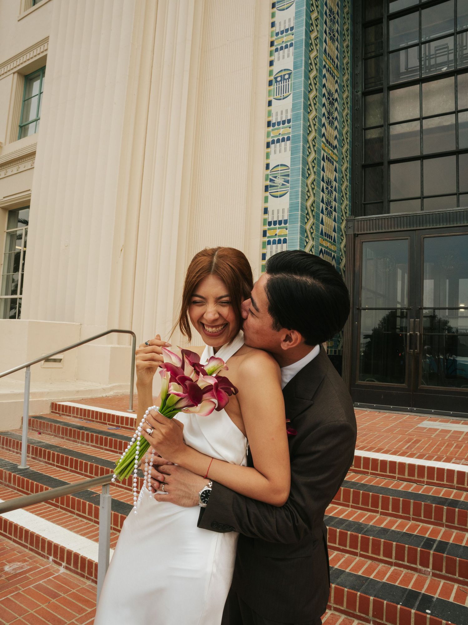A joyful bride holding a pink floral bouquet laughs as her groom kisses her cheek on brick steps.