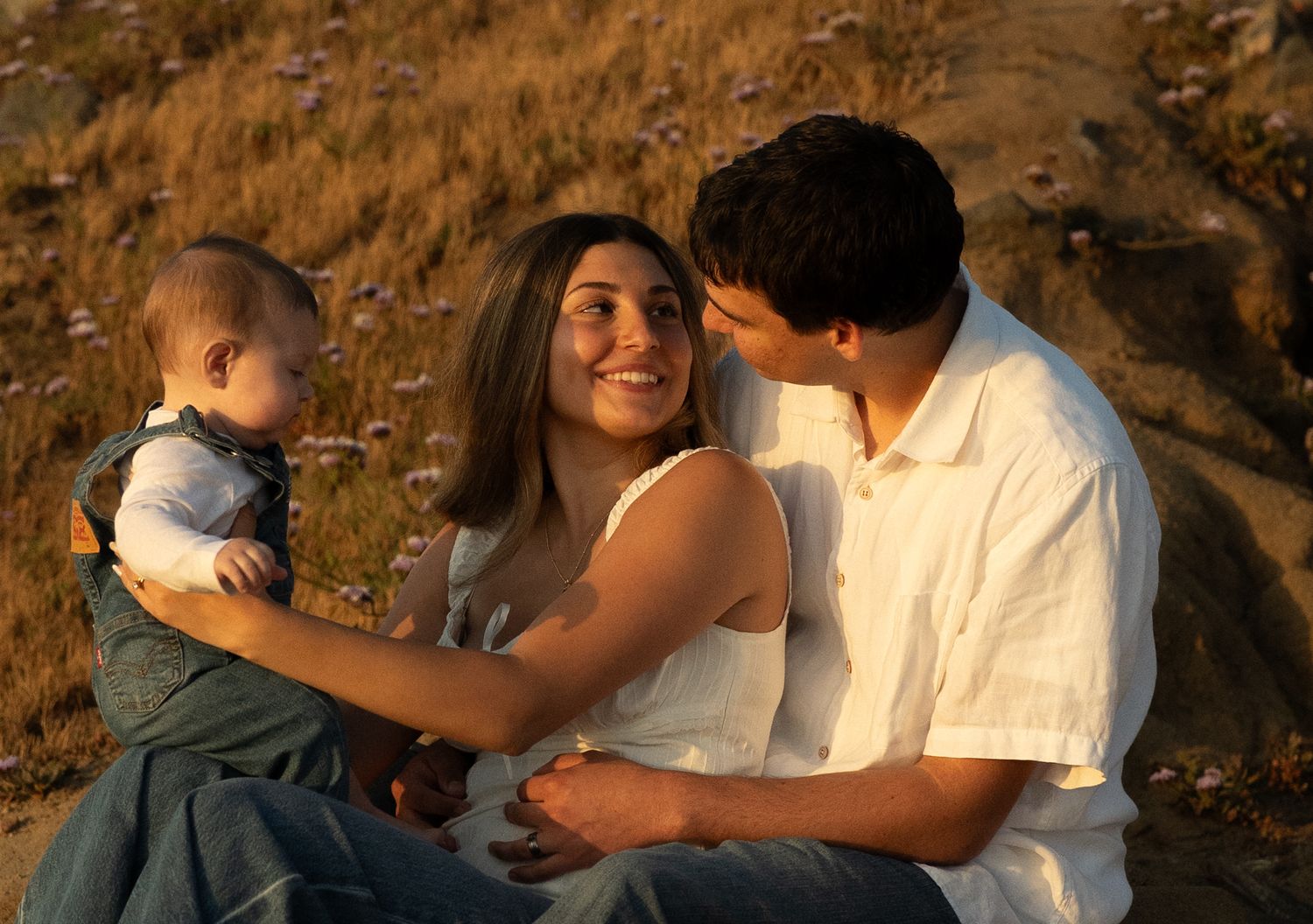 A young family sits outdoors at sunset, with a smiling mother holding a baby while a man leans in affectionately.
