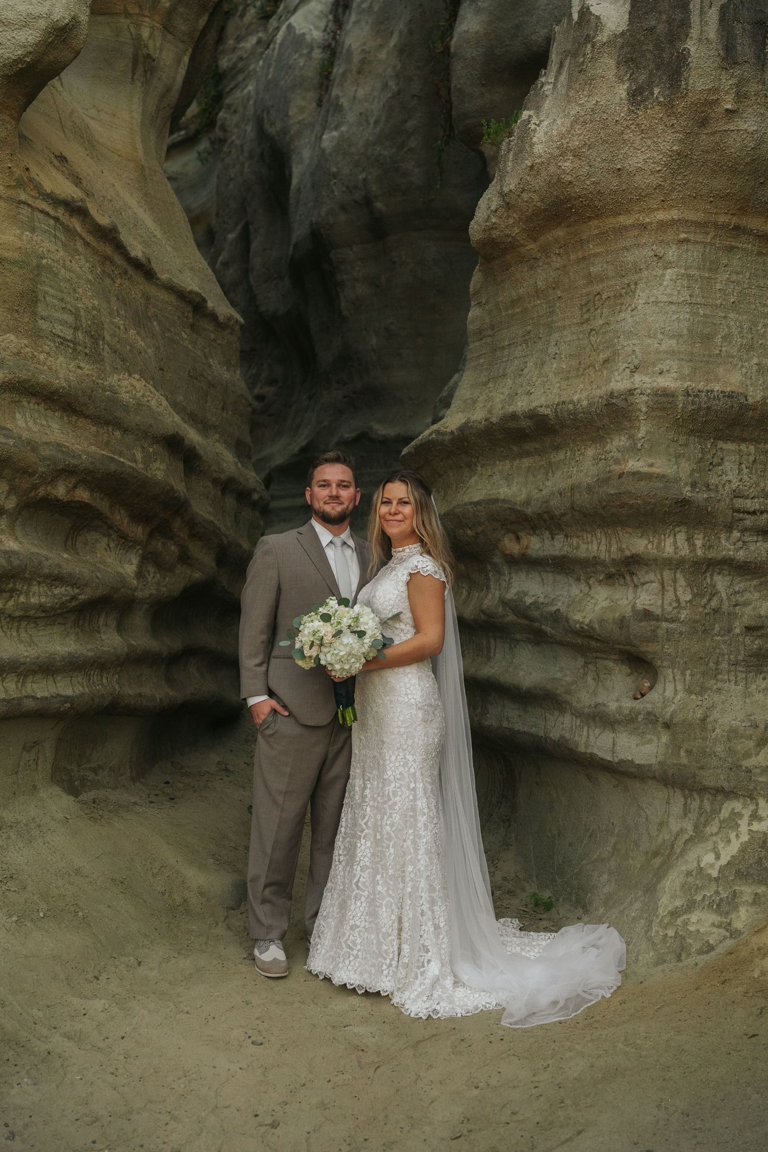 Bride in lace gown and groom in tan suit pose inside dramatic sandstone canyon with white bouquet.