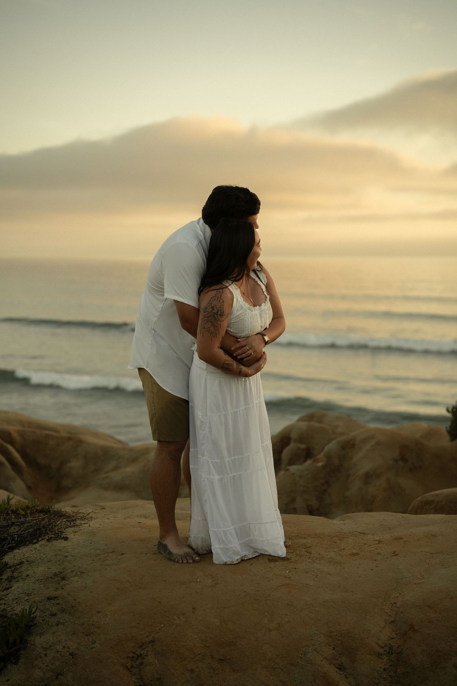 Couple standing on beach rocks at golden sunset, man embracing woman from behind overlooking the ocean.