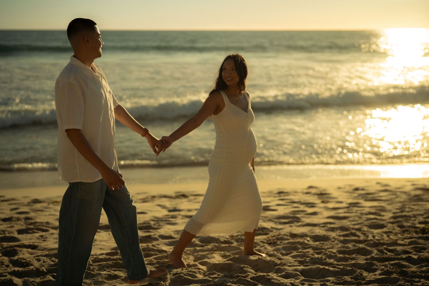 Pregnant couple holds hands walking on sandy beach at golden sunset.