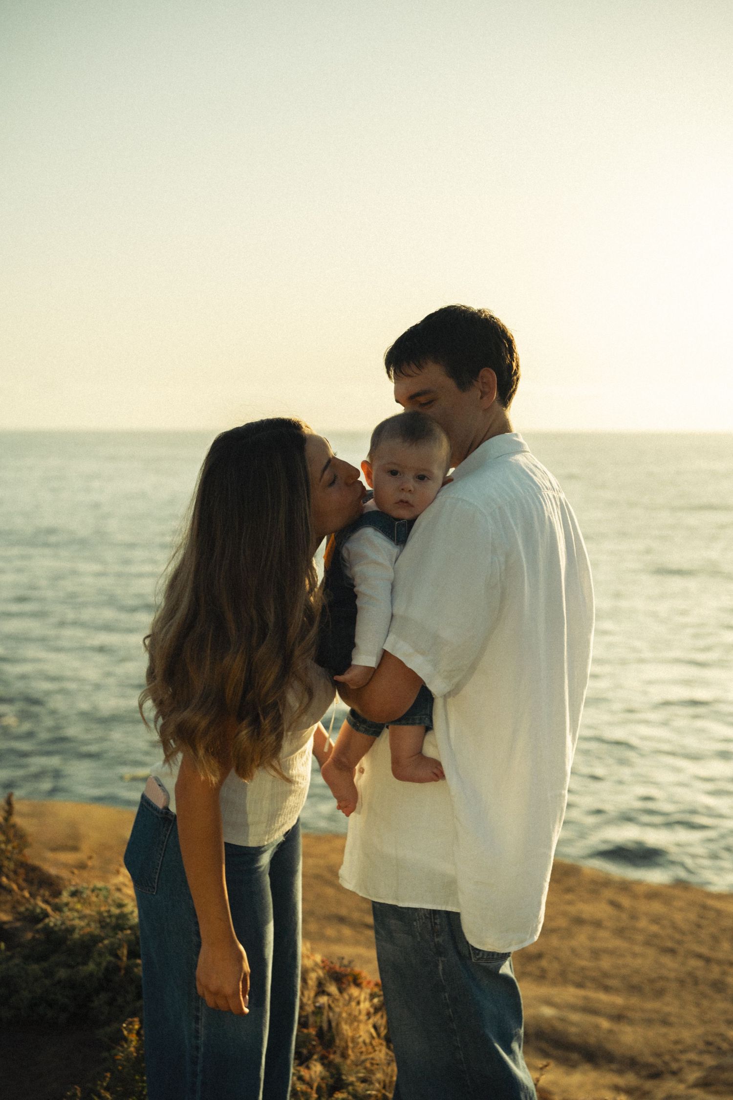 A young family shares a tender moment at sunset by the ocean, with parents kissing their baby.