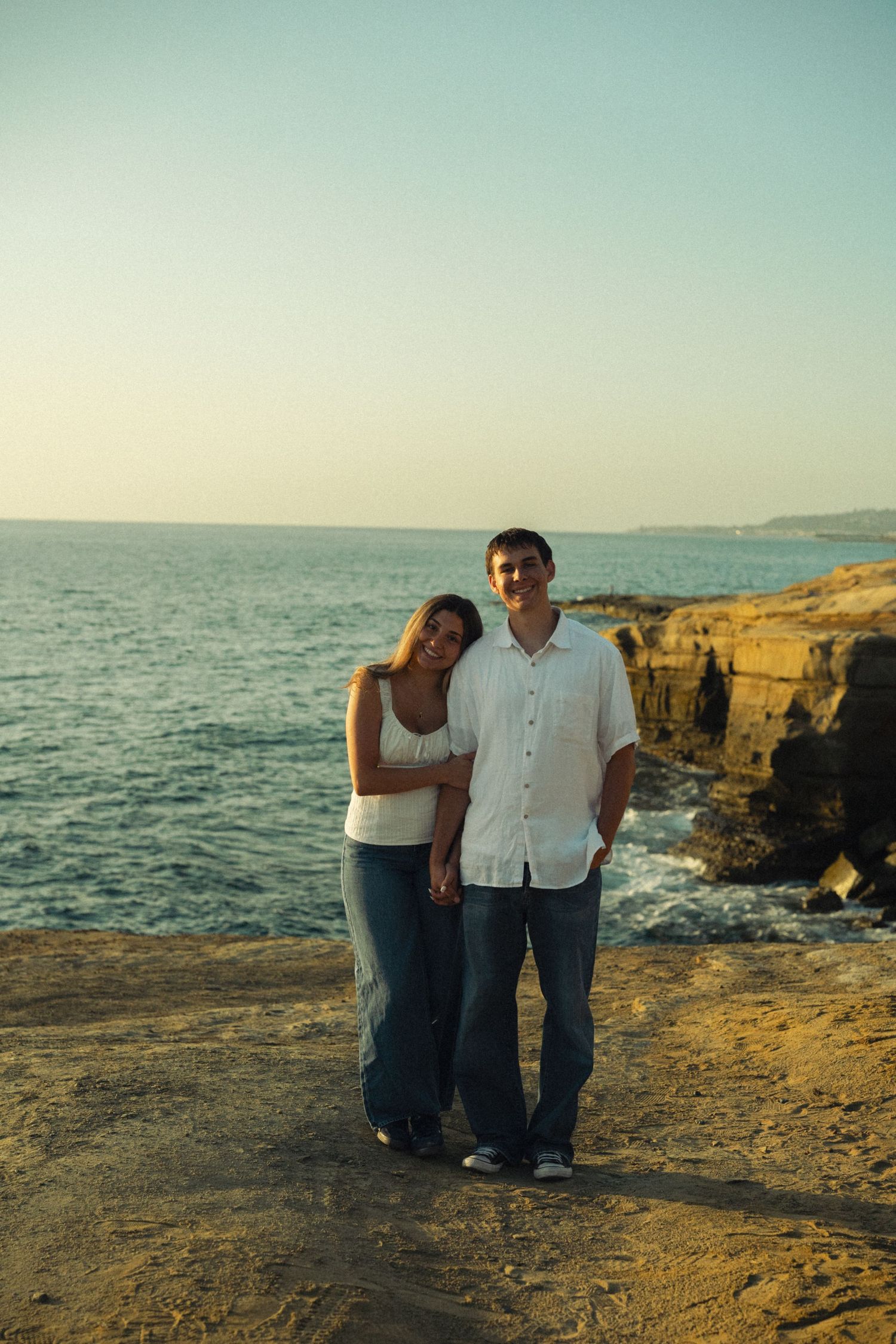 Couple posing together on rocky seaside cliff with ocean waves at golden sunset.