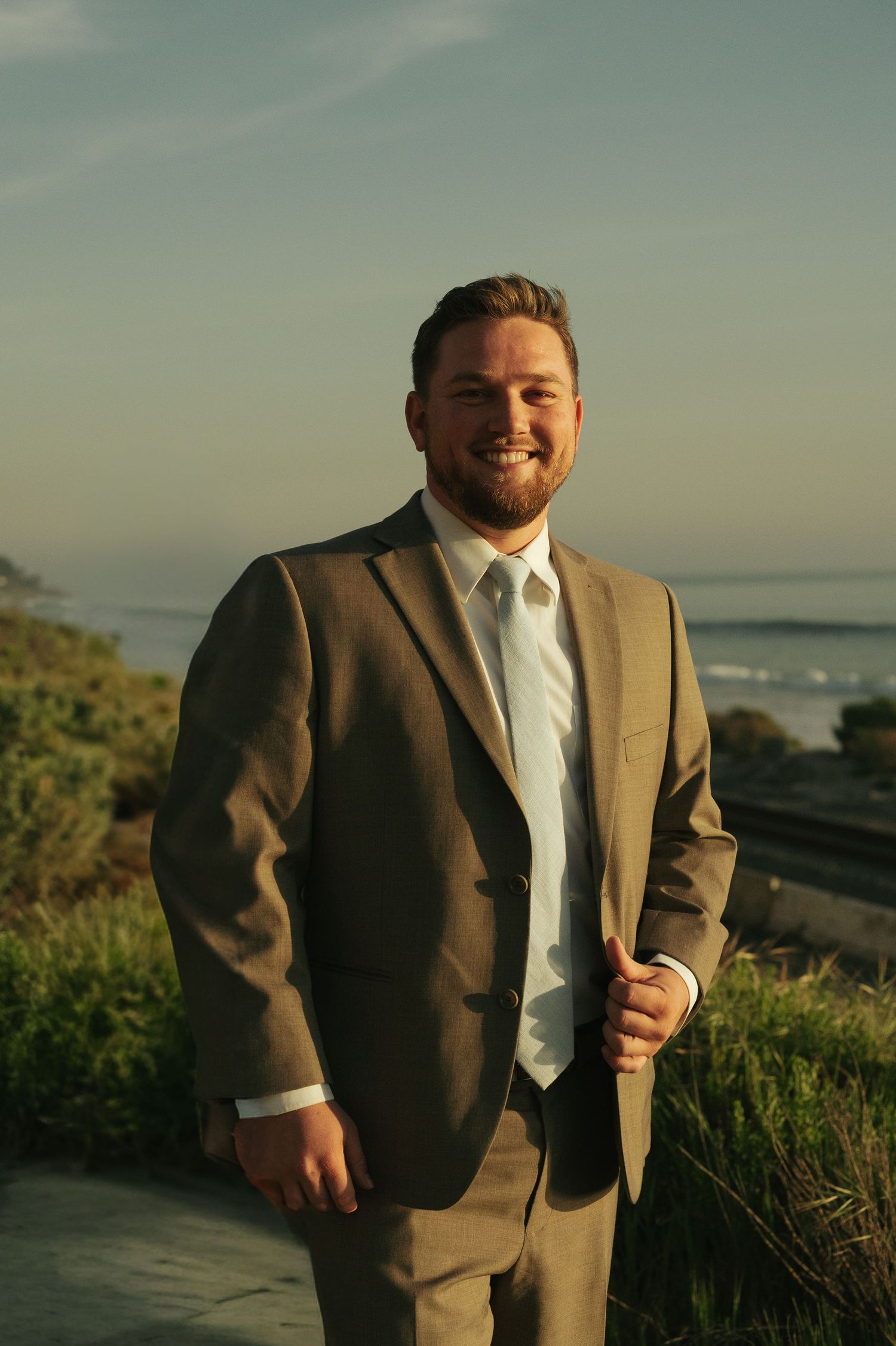 Smiling groom in a tan suit and tie poses outdoors near a coastal overlook at golden hour.