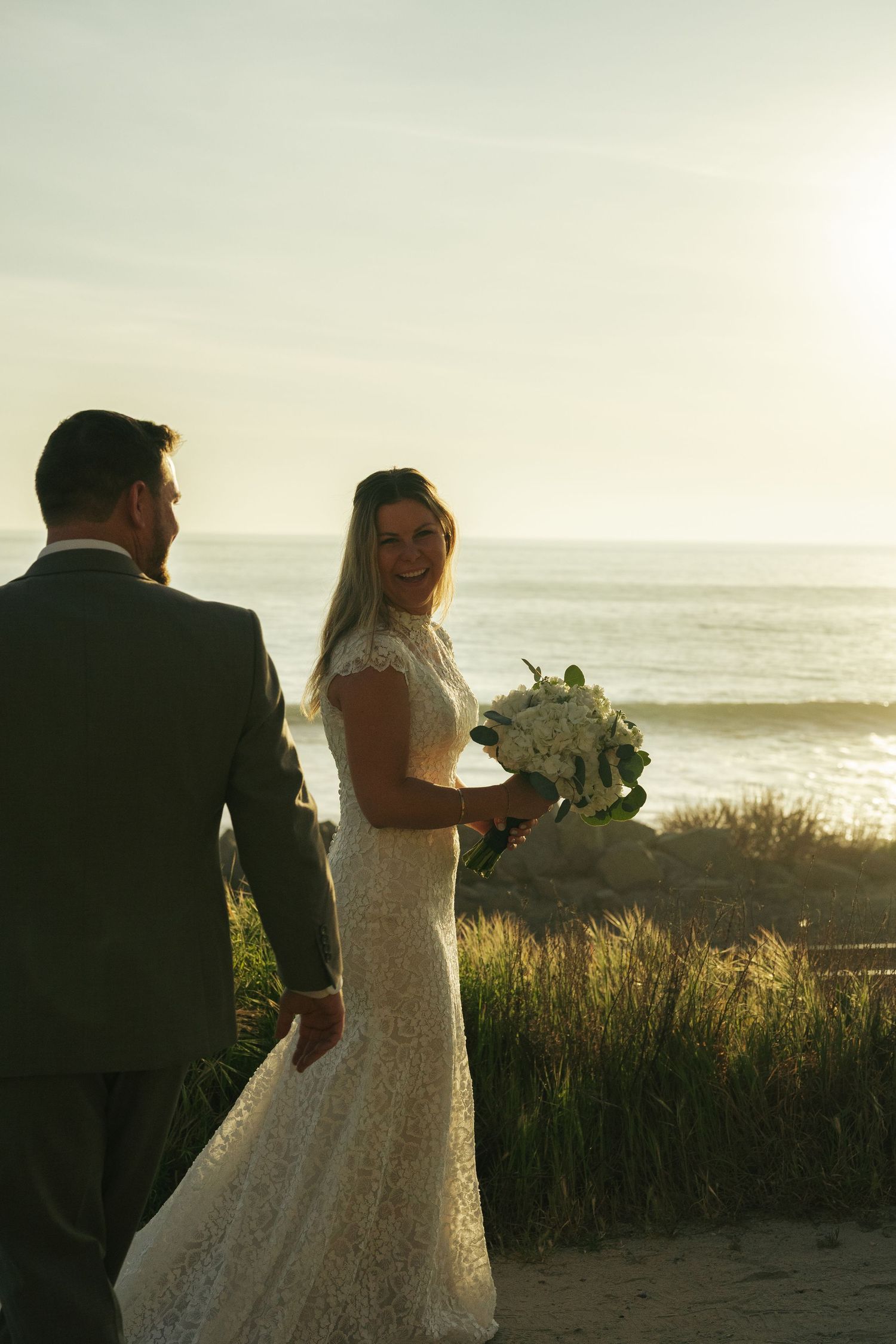 Smiling bride in lace wedding dress holding white bouquet walks with groom at golden hour beach sunset.