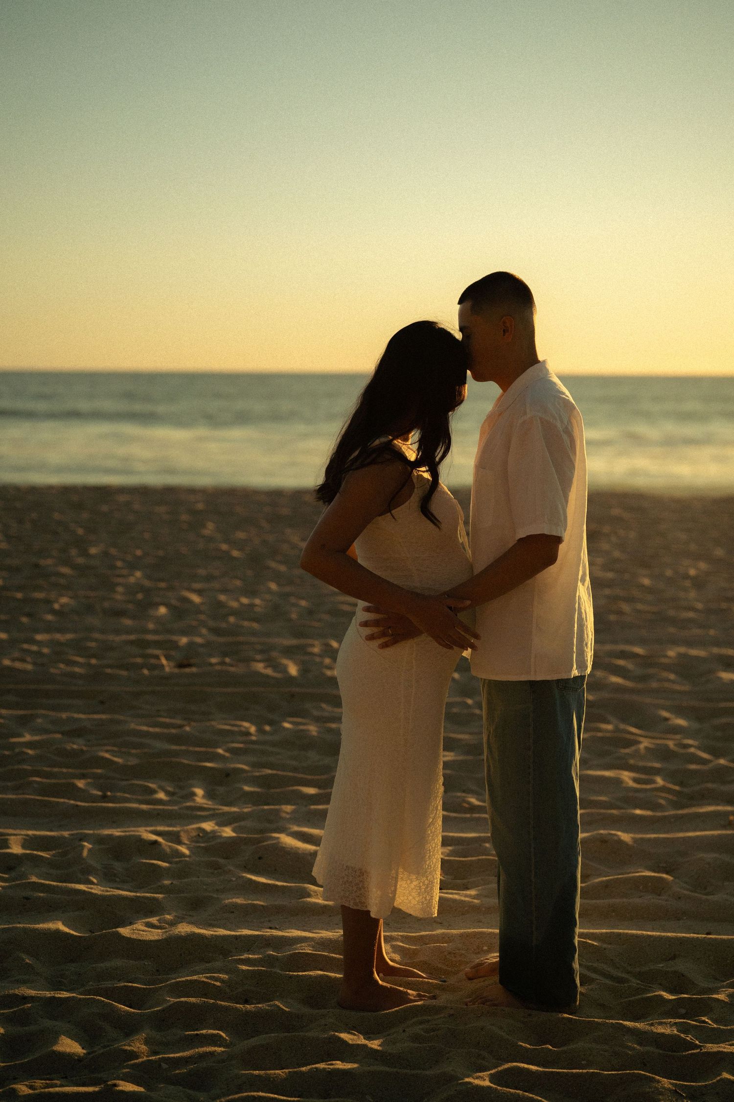 Silhouette of pregnant couple embracing on beach at sunset, man kissing womans forehead near ocean waves.